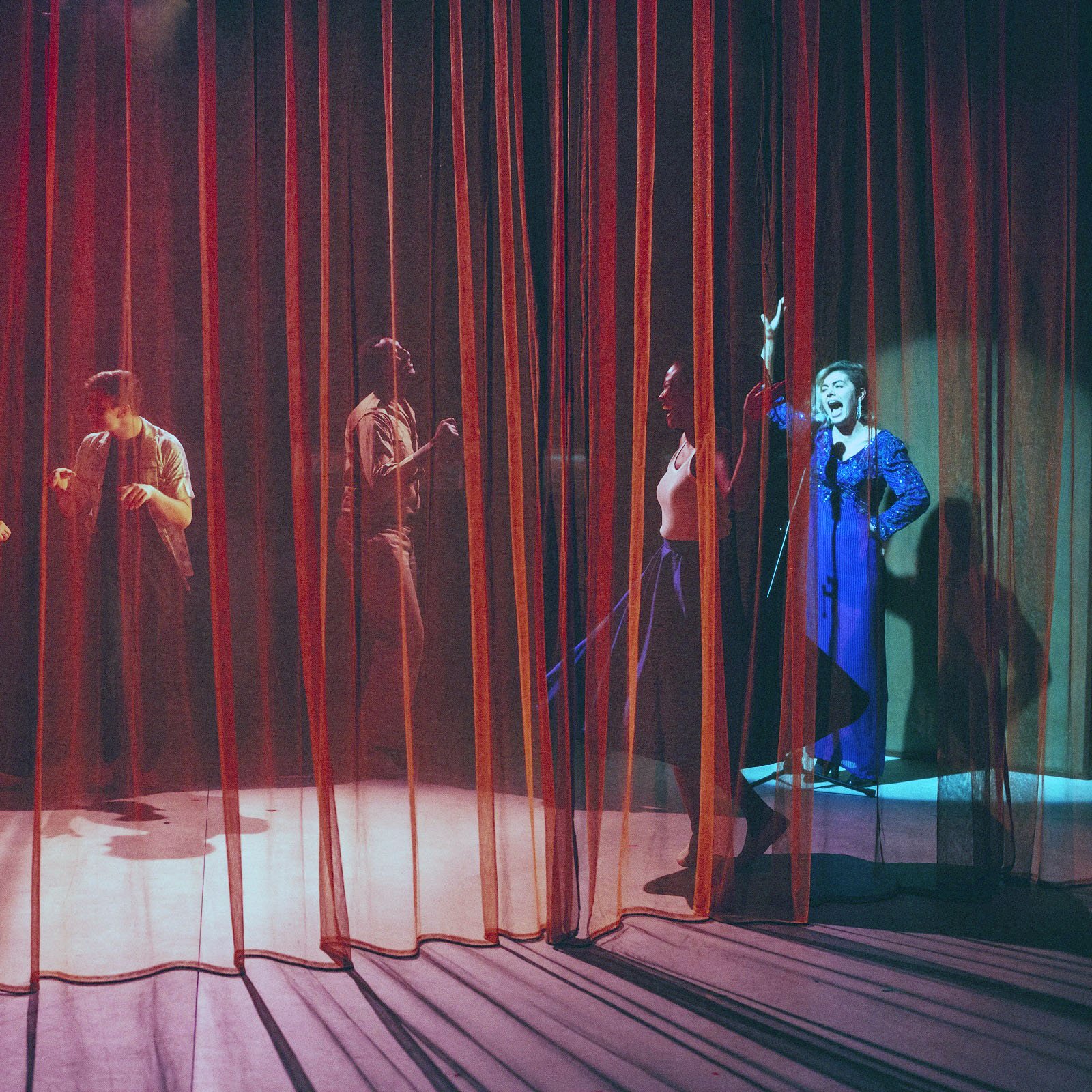 L'image montre une scène de théâtre avec des rideaux en velours rouge et orange qui créent une atmosphère intimiste. On aperçoit plusieurs silhouettes de personnes derrière les rideaux, engagées dans des activités qui semblent dynamiques et expressives. L'un des personnages, visible à droite, porte un costume bleu et semble gesticuler avec enthousiasme. Les jeux de lumière entre les rideaux ajoutent une ambiance dramatique à la scène, accentuant les mouvements des acteurs.