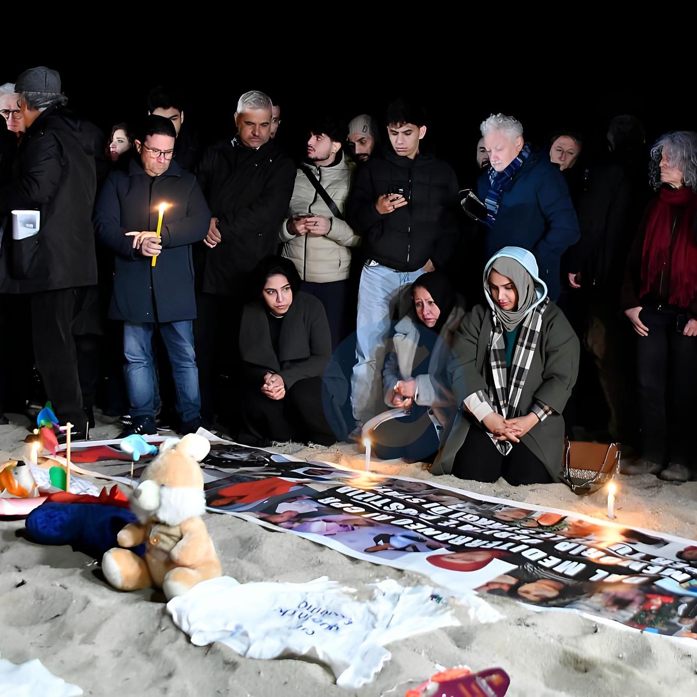 Un gruppo di persone in cerchio, con candele accese e striscioni, su una spiaggia al buio.