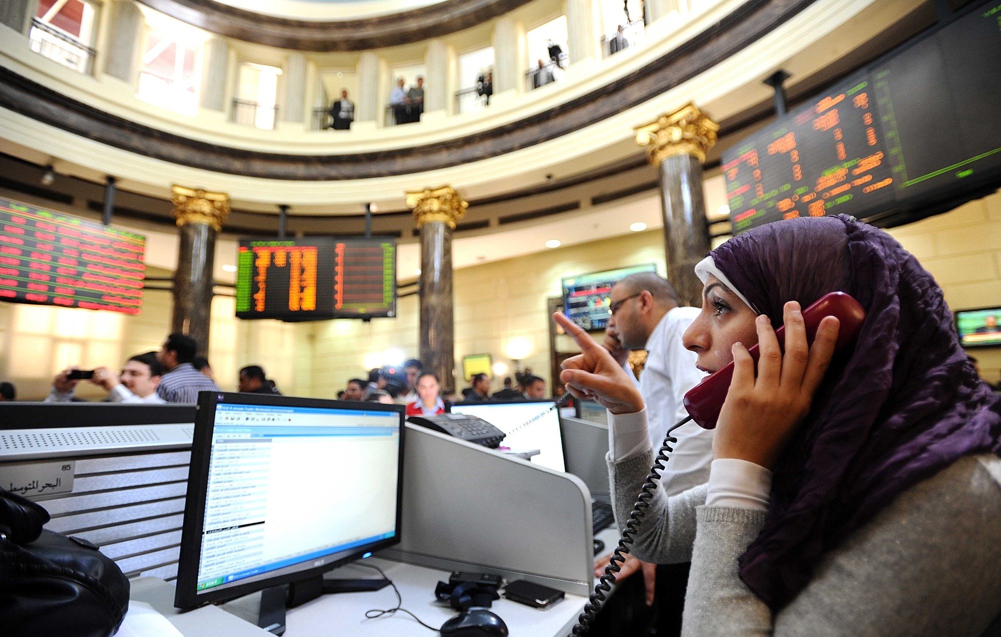 تظهر الصورة مشهداً من داخل سوق الأسهم، حيث يوجد عدد من الأشخاص يتعاملون مع شاشات تعرض الأسعار والبيانات المالية. في المقدمة، توجد امرأة ترتدي حجاباً، وهي تتحدث عبر الهاتف. يبدو أنها مشغولة بالعمل، حيث تشير بإحدى يديها. الخلفية تحتوي على عدة أشخاص آخرين وشاشات تعرض معلومات السوق. الأجواء توحي بالجدية والنشاط المالي.