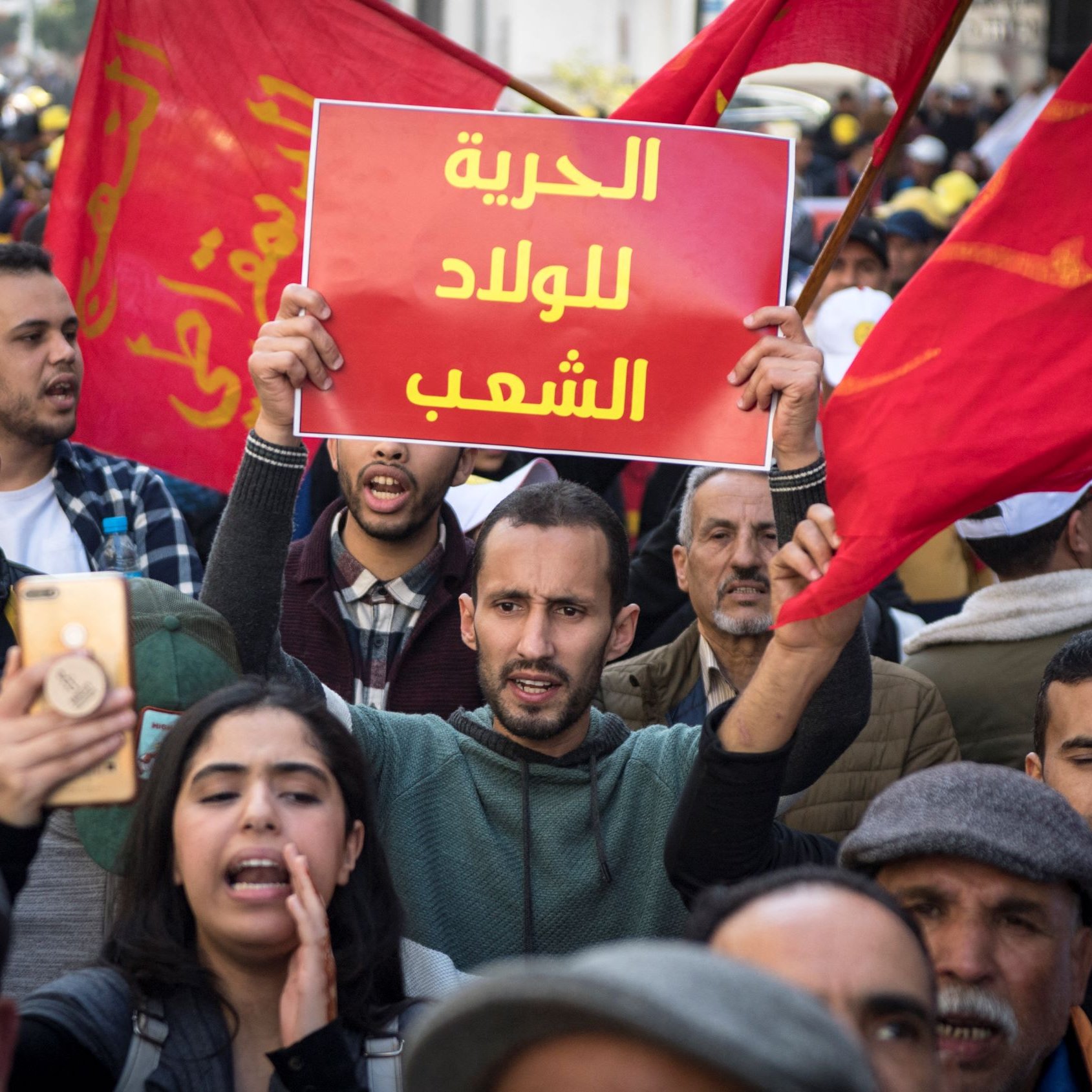 L'image montre une foule de manifestants, brandissant des drapeaux rouges. Au centre, un homme tient une pancarte avec le texte en arabe, tandis que d'autres personnes autour de lui semblent engagées dans la manifestation. On peut ressentir une atmosphère de protestation avec des expressions de détermination sur les visages des participants.