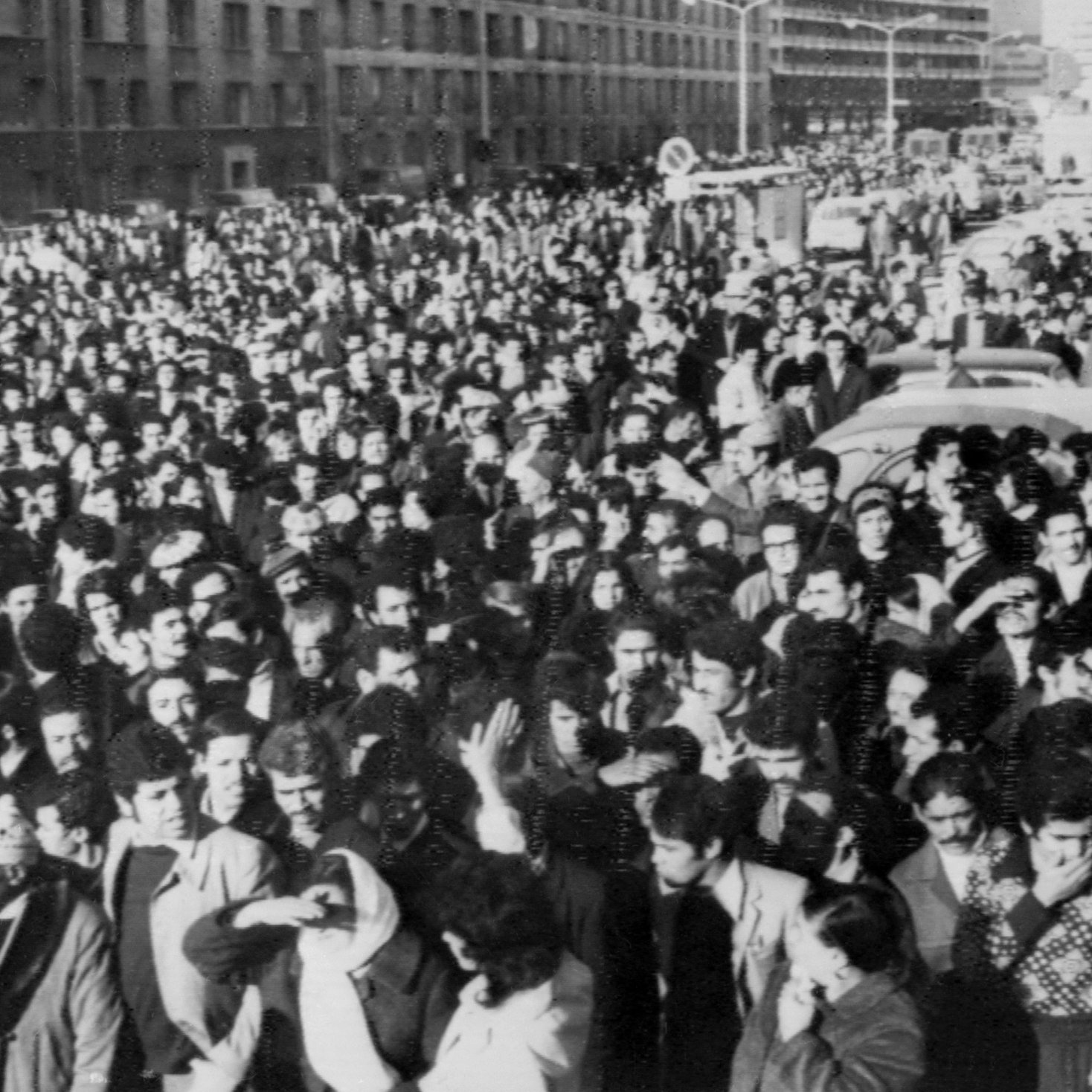 L'image montre une grande foule rassemblée dans une rue. Les personnes semblent se tenir très proches les unes des autres, créant une masse compacte. On peut apercevoir des bâtiments en arrière-plan, ainsi que des véhicules, ce qui indique que cette scène se déroule dans un milieu urbain. L'atmosphère semble intense et collective, suggérant un rassemblement pour une cause ou un événement significatif. Les vêtements des personnes, sombres et variés, donnent une impression de l'époque.
