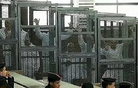 L'image montre un tribunal avec des accusés placés derrière des cages en métal ou des enclos. On aperçoit des personnes à l'intérieur de ces enclos, certaines levant la main. Il y a également des membres des forces de l'ordre en uniforme, observant la scène. L'environnement semble austère, et la lumière naturelle pénètre par des fenêtres en hauteur.