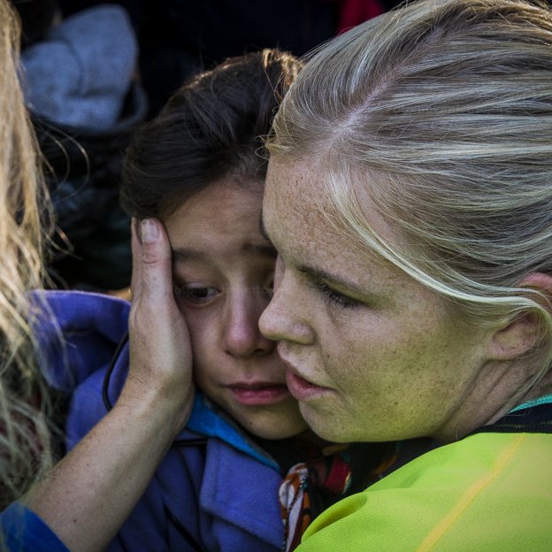 L'image montre une scène émotive où une femme, vêtue de vêtements de couleur vive, réconforte une petite fille. La femme semble douce et protectrice, tenant délicatement le visage de l'enfant. La petite fille a l'air triste ou inquiète, et elle se tourne vers la femme pour chercher du réconfort. L'environnement semble être chargé d'émotion, avec des personnes présentes en arrière-plan. Cette interaction illustre un moment de soutien et de compassion.