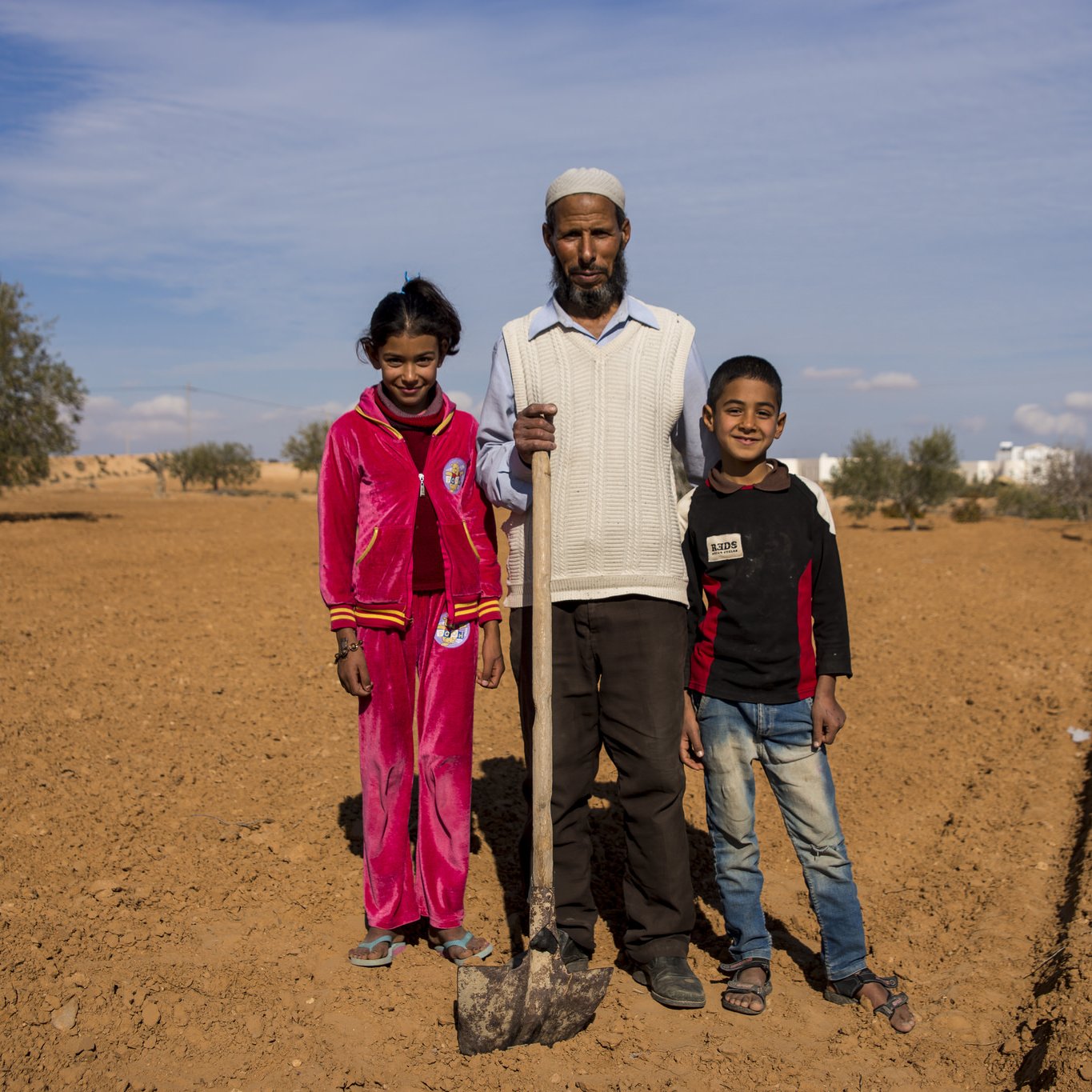 L'image montre un homme debout au milieu d'un paysage désertique, avec deux enfants près de lui, une fille et un garçon. L'homme tient une pelle et porte des vêtements simples. La fille est vêtue d'un ensemble rouge, tandis que le garçon porte un sweat-shirt noir avec des détails rouges. En arrière-plan, on peut voir quelques arbres éparpillés et un ciel bleu clair. Le sol est sec et nu, typique d'une région aride. L'ambiance semble conviviale, suggérant une journée de travail ou de récolte en famille.