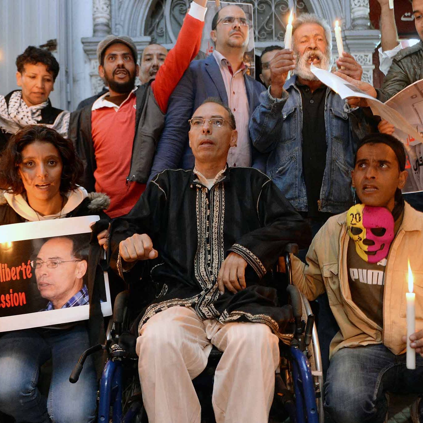 L'image montre un groupe de personnes rassemblées, tenant des bougies allumées. Au premier plan, un homme en fauteuil roulant est au centre, tenant une pancarte. Les autres personnes autour de lui semblent participer à une manifestation pacifique, certaines brandissant des affiches et d'autres affichant des expressions sérieuses. L'ambiance semblerait être axée sur la défense des droits et de la liberté d'expression.