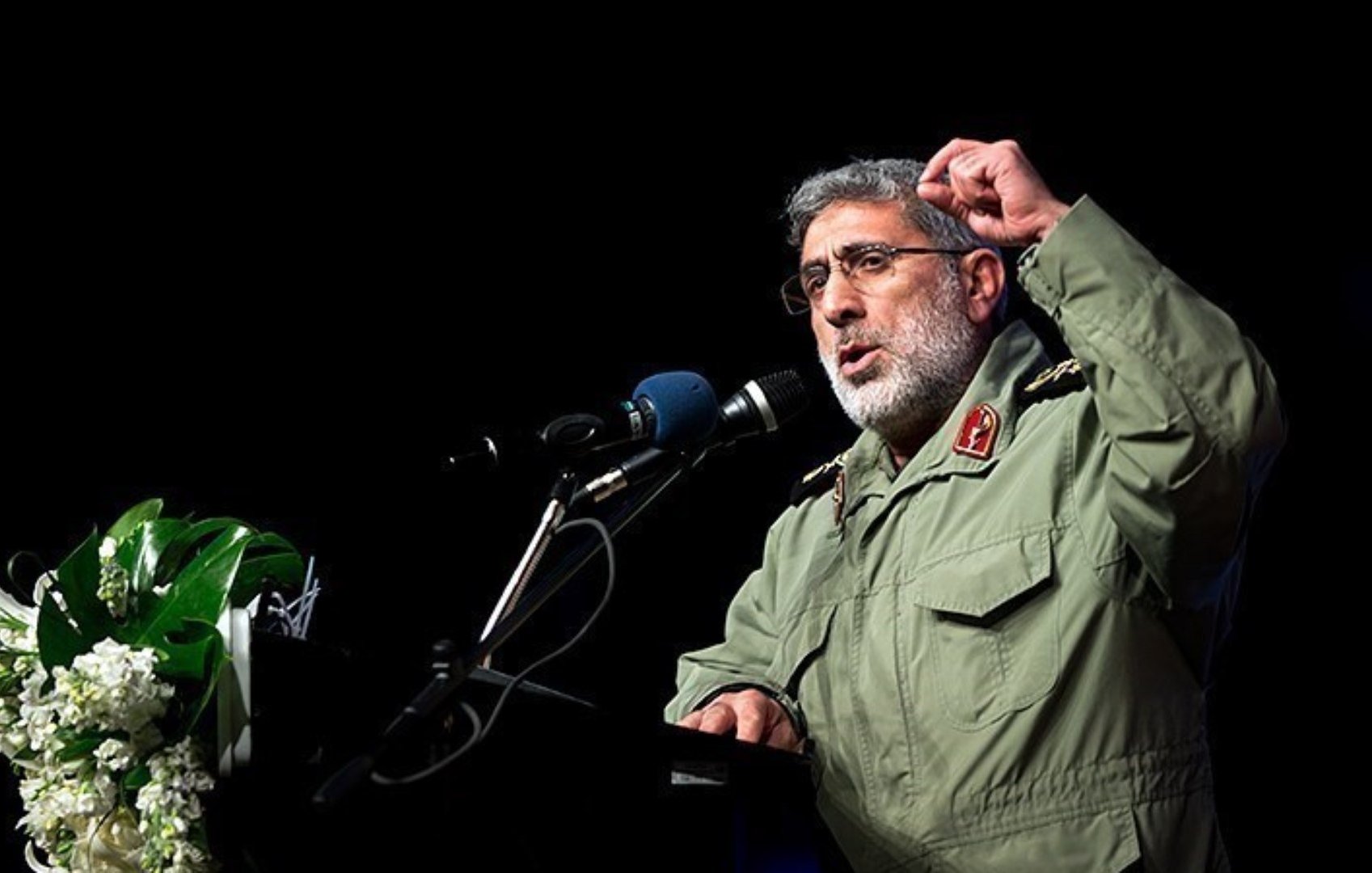 L'image montre un homme en uniforme militaire, debout devant un micro sur une scène. Il semble s'adresser à un public, probablement en train de faire un discours. Son expression est sérieuse, et il gesticule avec la main pour soutenir ses propos. Sur le podium, on peut voir des fleurs ou des plantes qui ajoutent une touche décorative à la scène.