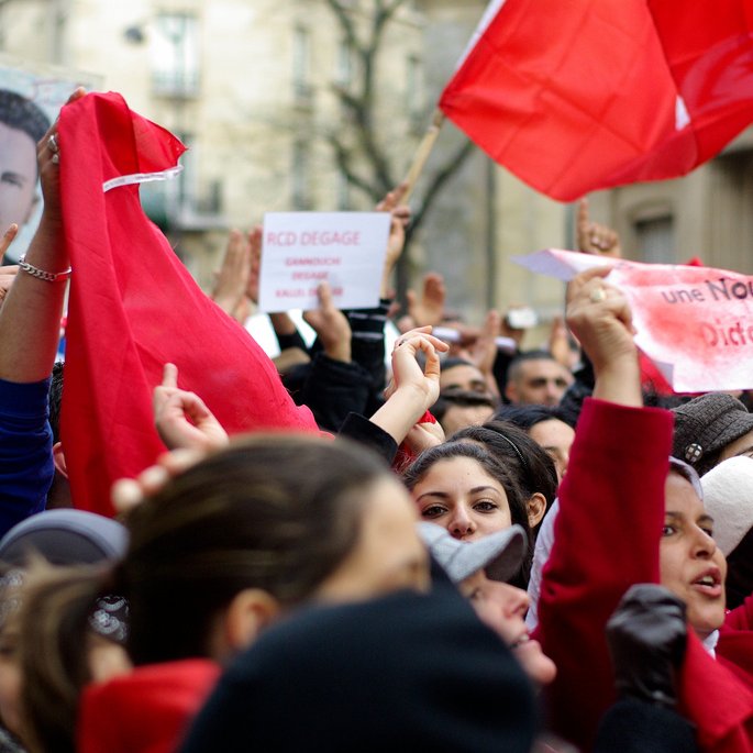 L'image montre une foule de manifestants rassemblés, brandissant des drapeaux rouges et des pancartes. Certains visages sont masqués, tandis que d'autres expriment des émotions intenses, comme la détermination ou la colère. On aperçoit aussi un portrait d'une figure emblématique en arrière-plan. Les slogans sur les pancartes semblent appeler au changement et à la liberté, indiquant un contexte de protestation politique. L'atmosphère est chargée d'énergie collective.
