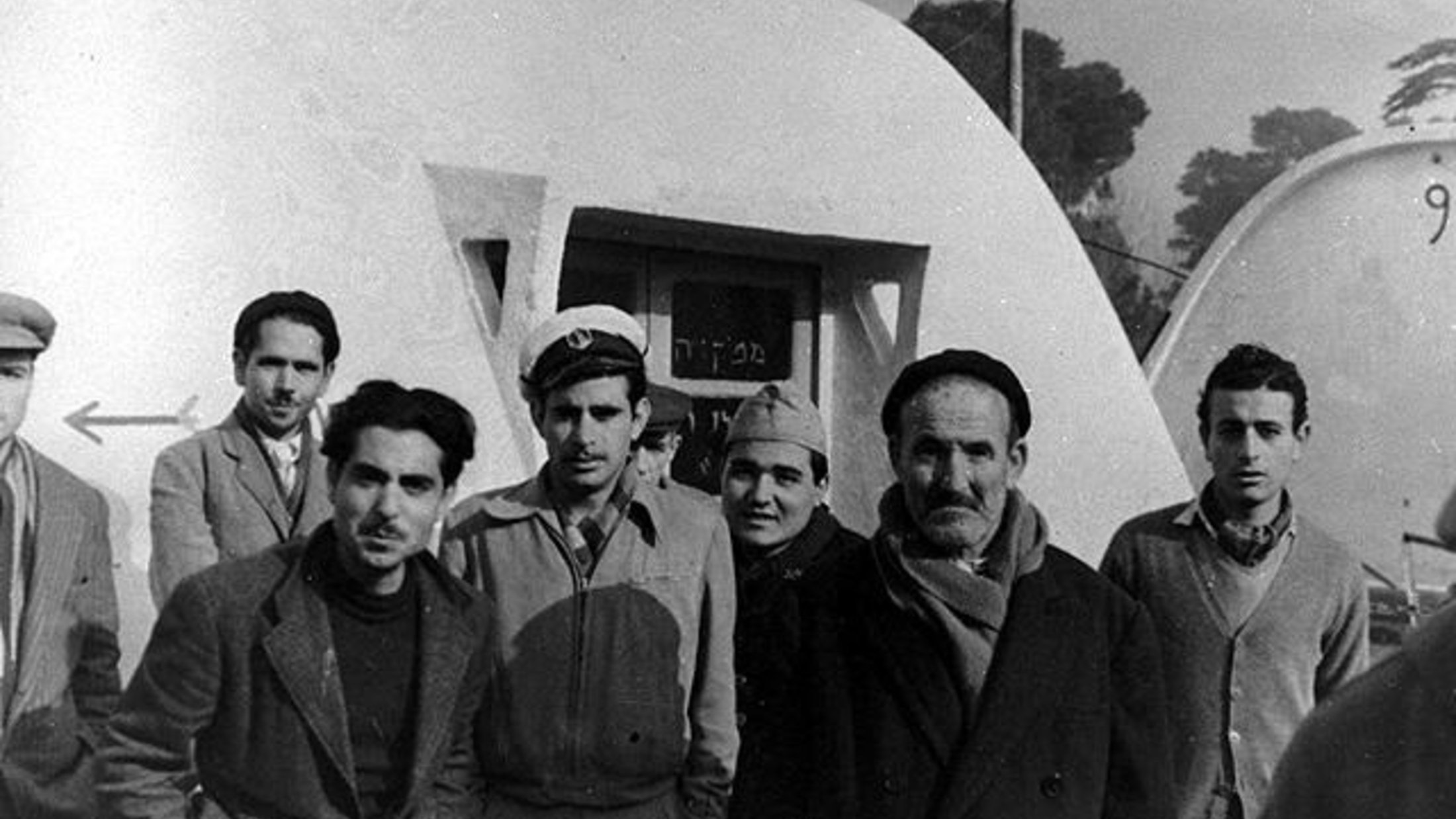 L'image montre un groupe de six hommes, debout près d'une structure blanche ressemblant à une maison ou une cabane. Ils sont habillés de manière variée, certains portant des chapeaux et des manteaux, ce qui suggère un temps frais. L'arrière-plan présente une autre structure similaire et des arbres flous. L'atmosphère semble décontractée, mais les expressions des hommes sont sérieuses.