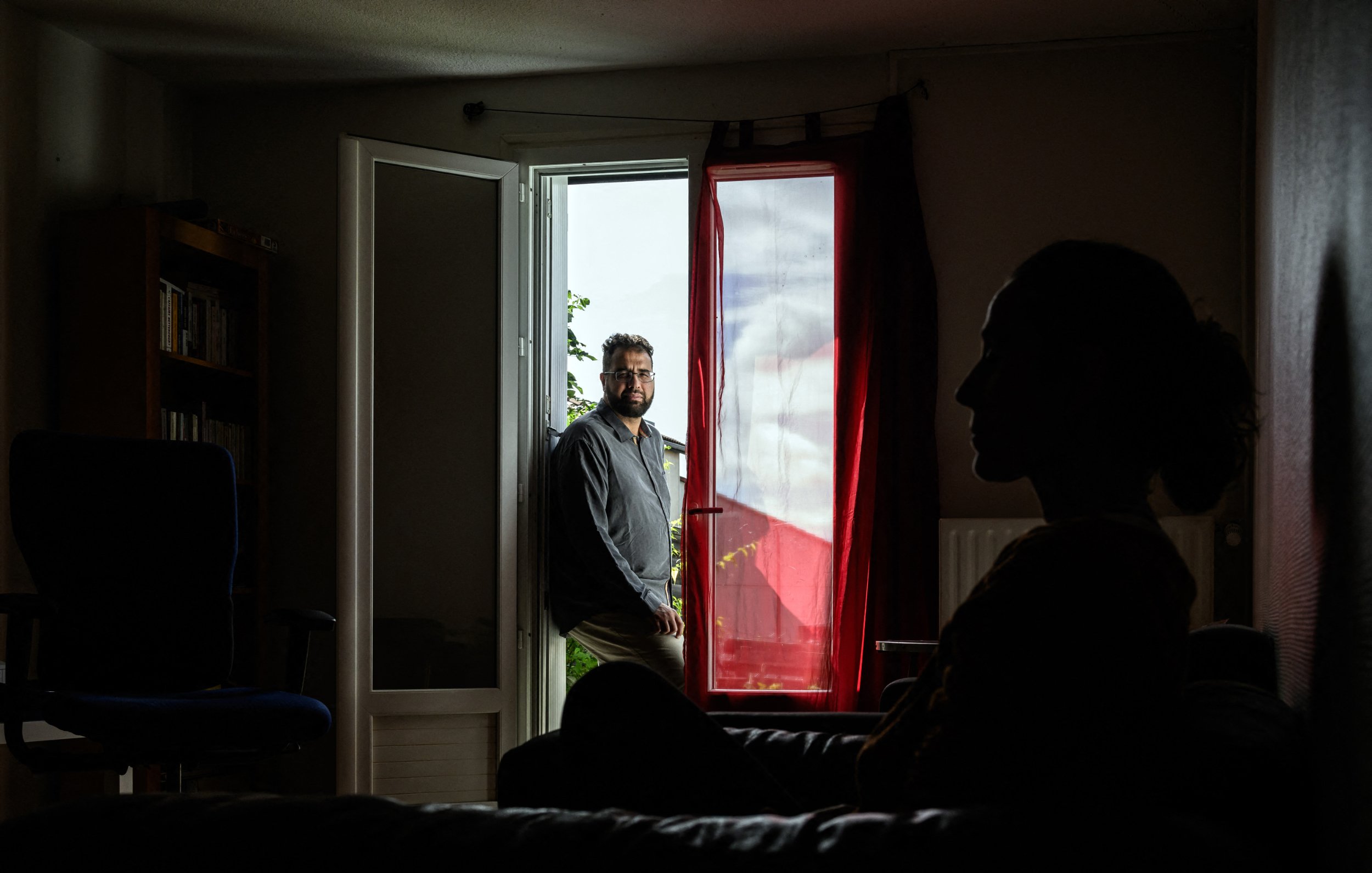 L'image montre un intérieur de maison ou d'appartement. Au premier plan, on voit la silhouette d'une personne assise sur un canapé, éclairée par une lumière douce, tandis qu'en arrière-plan, un homme est debout près d'une porte ouverte qui donne sur l'extérieur. La lumière entre par la fenêtre de façon à créer un contraste intéressant entre les deux personnages. Les éléments de la pièce, tels qu'un fauteuil et des étagères, ajoutent à l'ambiance chaleureuse et intime de la scène.