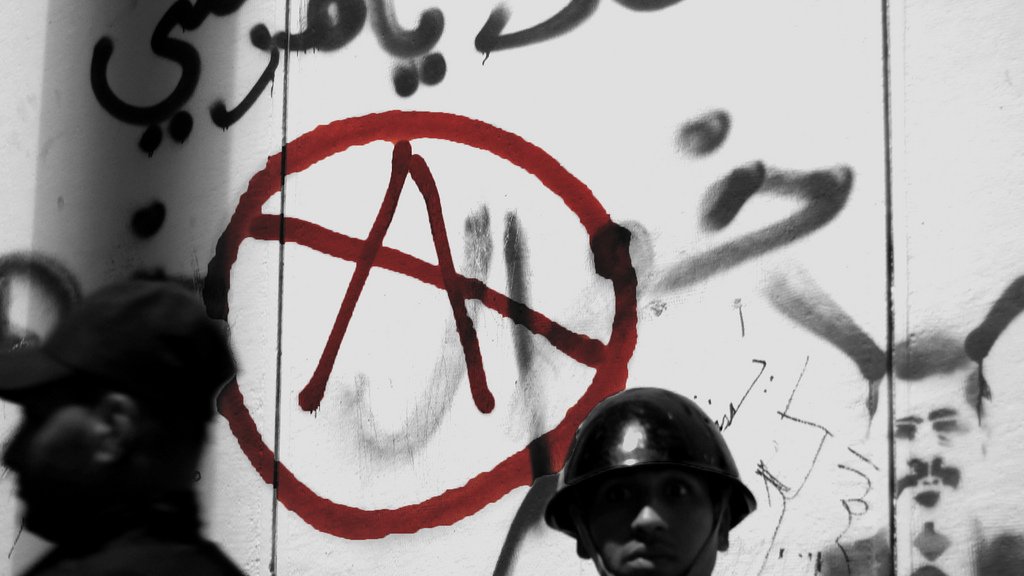 L'image présente un mur recouvert de graffitis, dont un symbole anarchiste stylisé en rouge, entouré d'un cercle. En arrière-plan, on voit deux hommes en uniforme, probablement des forces de l'ordre. L'un d'eux est au premier plan, portant un casque, avec une expression sérieuse. L'atmosphère de la photo suggère une tension sociale, accentuée par l'utilisation de couleurs sombres et le contraste avec le symbole rouge.