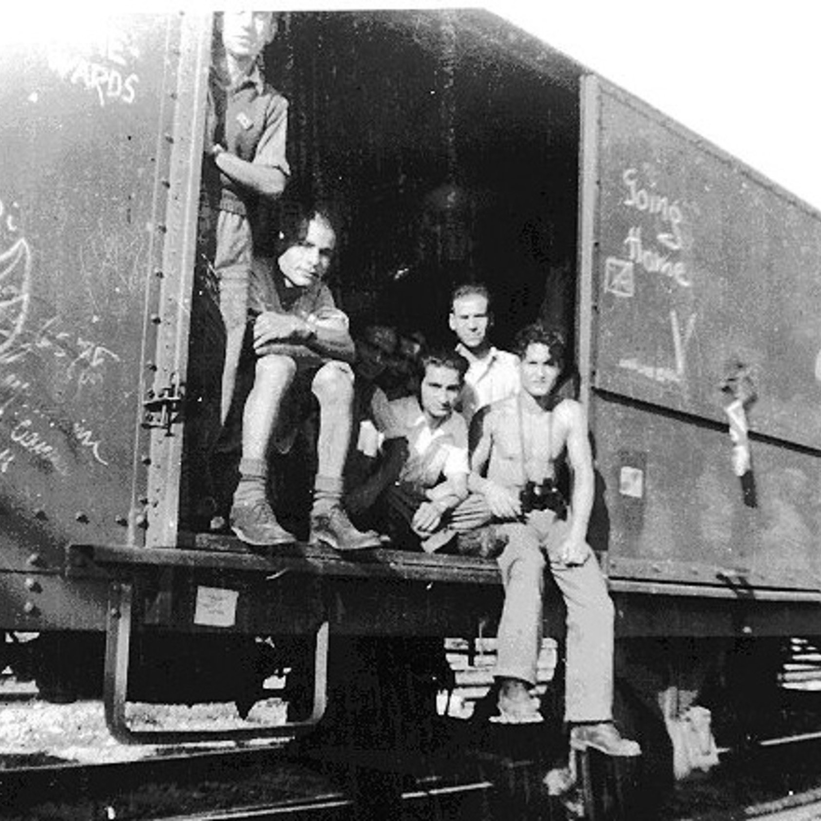 L'image montre un groupe de jeunes hommes assis et debout dans un wagon de train. Ils semblent se trouver dans un environnement rural ou industriel, avec des rails visibles au premier plan. Les murs du wagon sont couverts de graffitis, et l'ambiance générale paraît nostalgique. Les expressions des jeunes varient, allant de la réflexion à l'insouciance. L'image est en noir et blanc, ce qui lui donne un aspect vintage.
