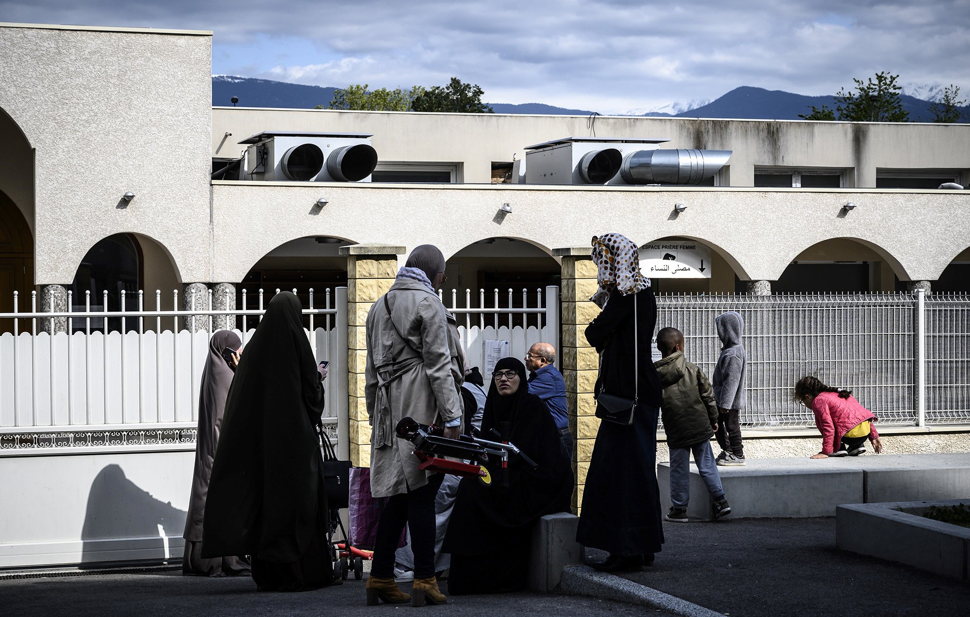 L'image montre un groupe de personnes à l'extérieur d'un bâtiment. Certaines femmes portent des vêtements longs et des foulards, tandis que d'autres semblent habillées plus décontractées. On aperçoit également des enfants jouant dans l'arrière-plan. Le ciel est partiellement nuageux et des montagnes se dessinent au loin. L'ambiance semble conviviale, avec des échanges entre les personnes présentes.
