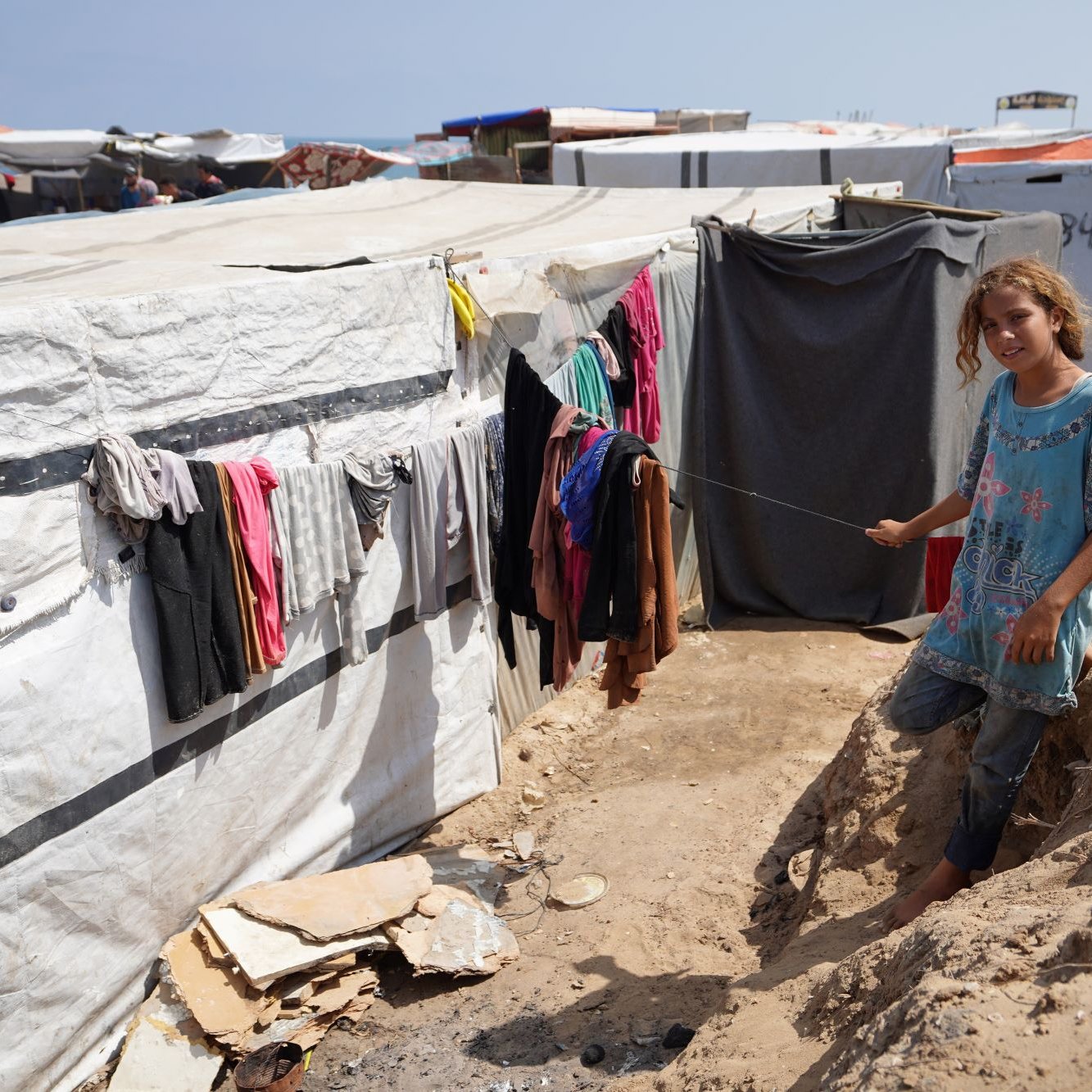 L'image montre un camp de tentes ou de structures temporaires. À gauche, des vêtements sont accrochés à une corde à linge, tandis qu'une jeune fille se trouve au premier plan, tenant une sorte de bâton. Elle porte une tunique et semble se tenir sur un chemin étroit entre les tentes. Le décor est marqué par des bords de sol en terre et la structure du camp qui se développe à l'arrière-plan. L'atmosphère semble celle d'un environnement difficile, lié à des conditions de vie précaires.