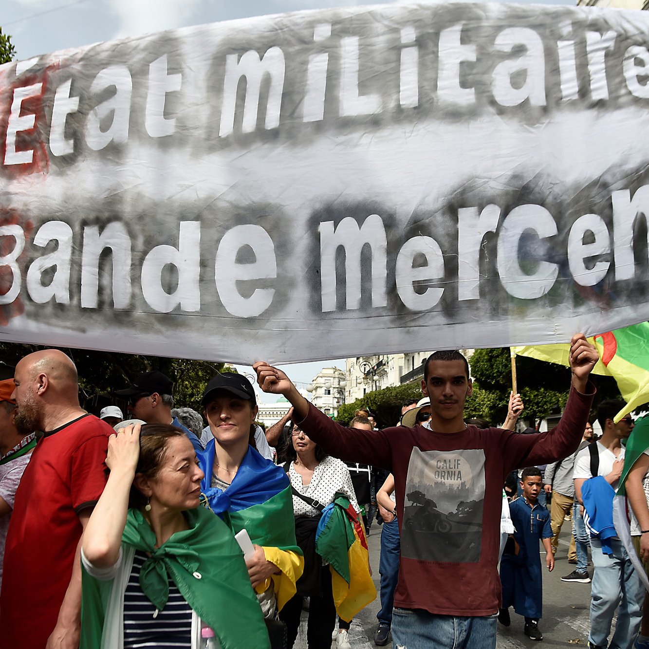 الصورة تُظهر مجموعة من الناس في مسيرة أو احتجاج، حيث يحمل أحد المشاركين لافتة كبيرة مكتوب عليها "لا للدولة العسكرية ولا للفرقة المرتزقة". يبدو أن المشاركين يعبرون عن مطالب سياسية أو اجتماعية، كما يوضح تنوع الحضور من حيث الأعمار والجنسيات. الأجواء تعكس حماس المتظاهرين وتضامنهم.