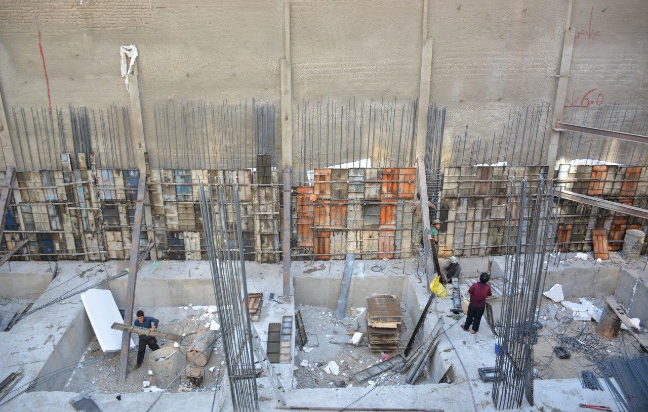 L'image montre un chantier de construction. On peut y voir des ouvriers qui travaillent dans une excavation, entourés de barres d'acier et de matériaux de construction. Les murs en béton sont partiellement visibles, avec des panneaux de coffrage en bois et en métal disposés verticalement. L'environnement semble être en cours de développement, avec une variété d'outils et de matériaux éparpillés autour de l'espace de travail.