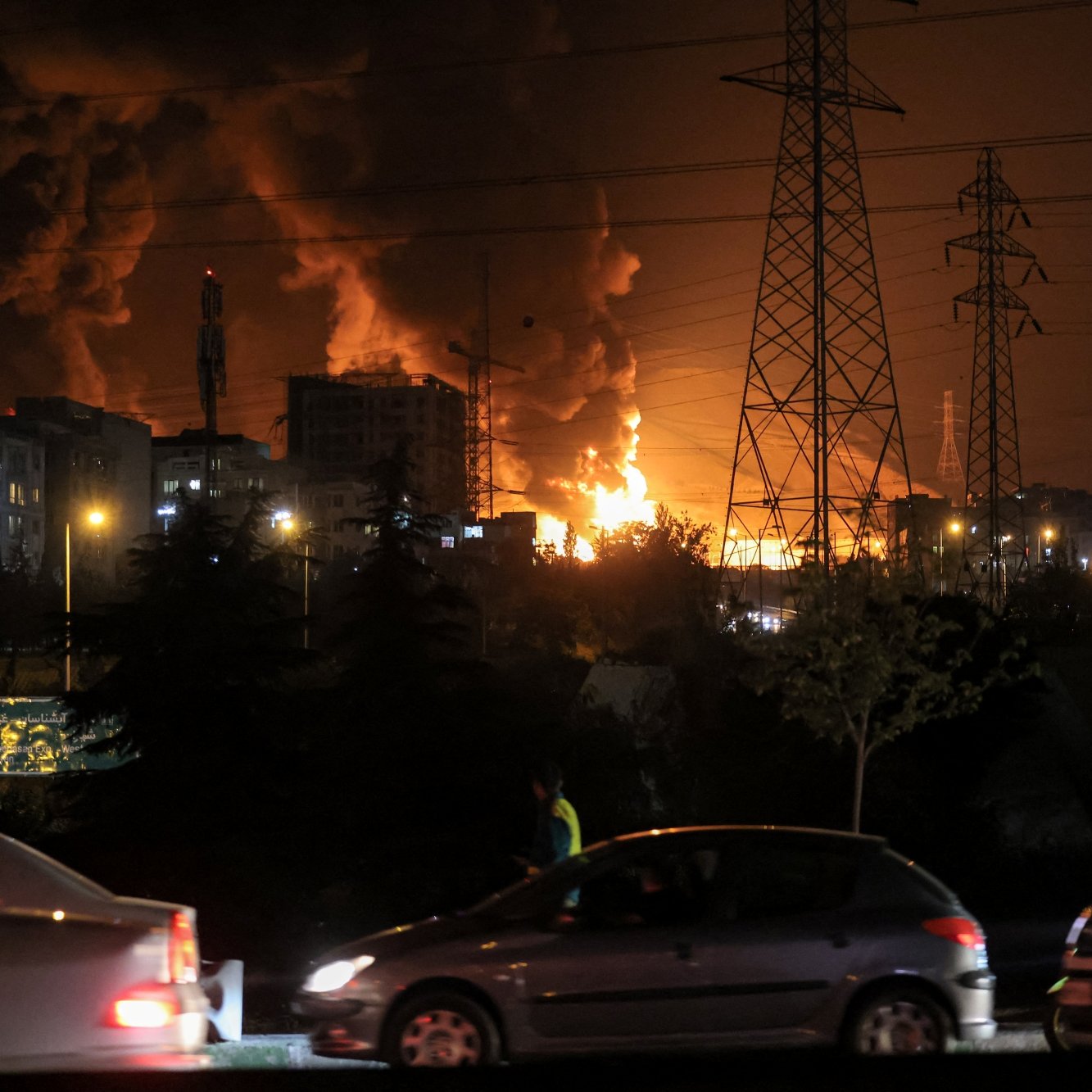 تصویری تاریک با آتش و دود بزرگ، ساختمان‌ها و تیرک‌های برق در پس‌زمینه، و خودروهای در حال حرکت.