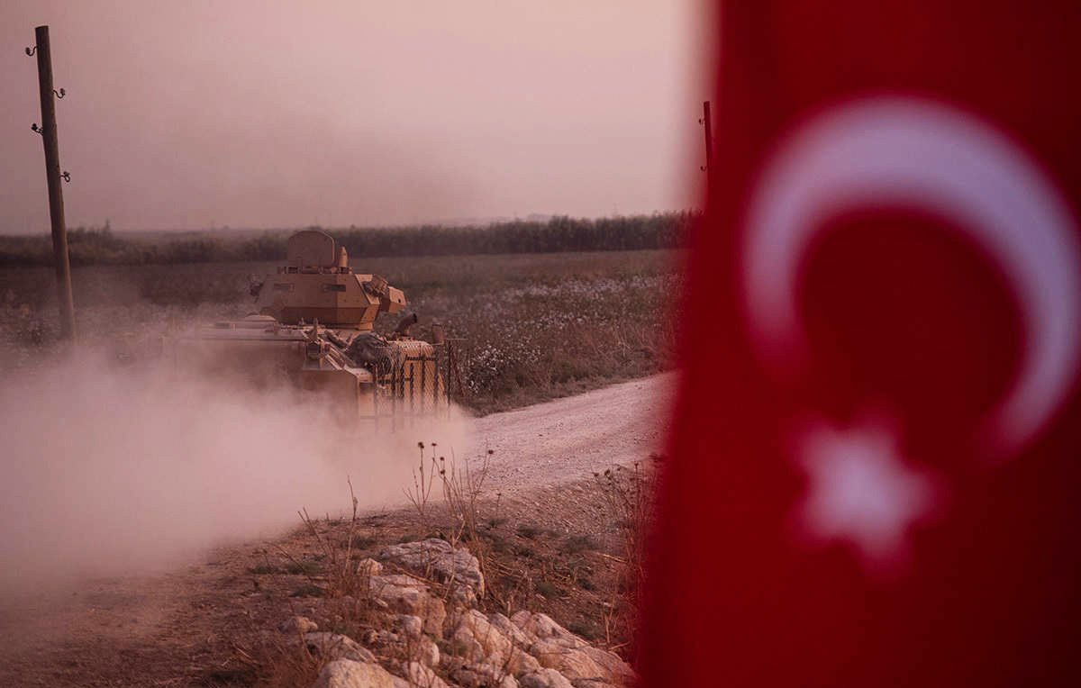 The image depicts a military tank moving along a dirt road, creating a cloud of dust. In the foreground, part of a Turkish flag is visible, featuring a white star and crescent on a red background. The landscape appears to be rural, possibly with fields or open land in the background, and the lighting suggests either dawn or dusk, giving a somewhat dramatic atmosphere to the scene.