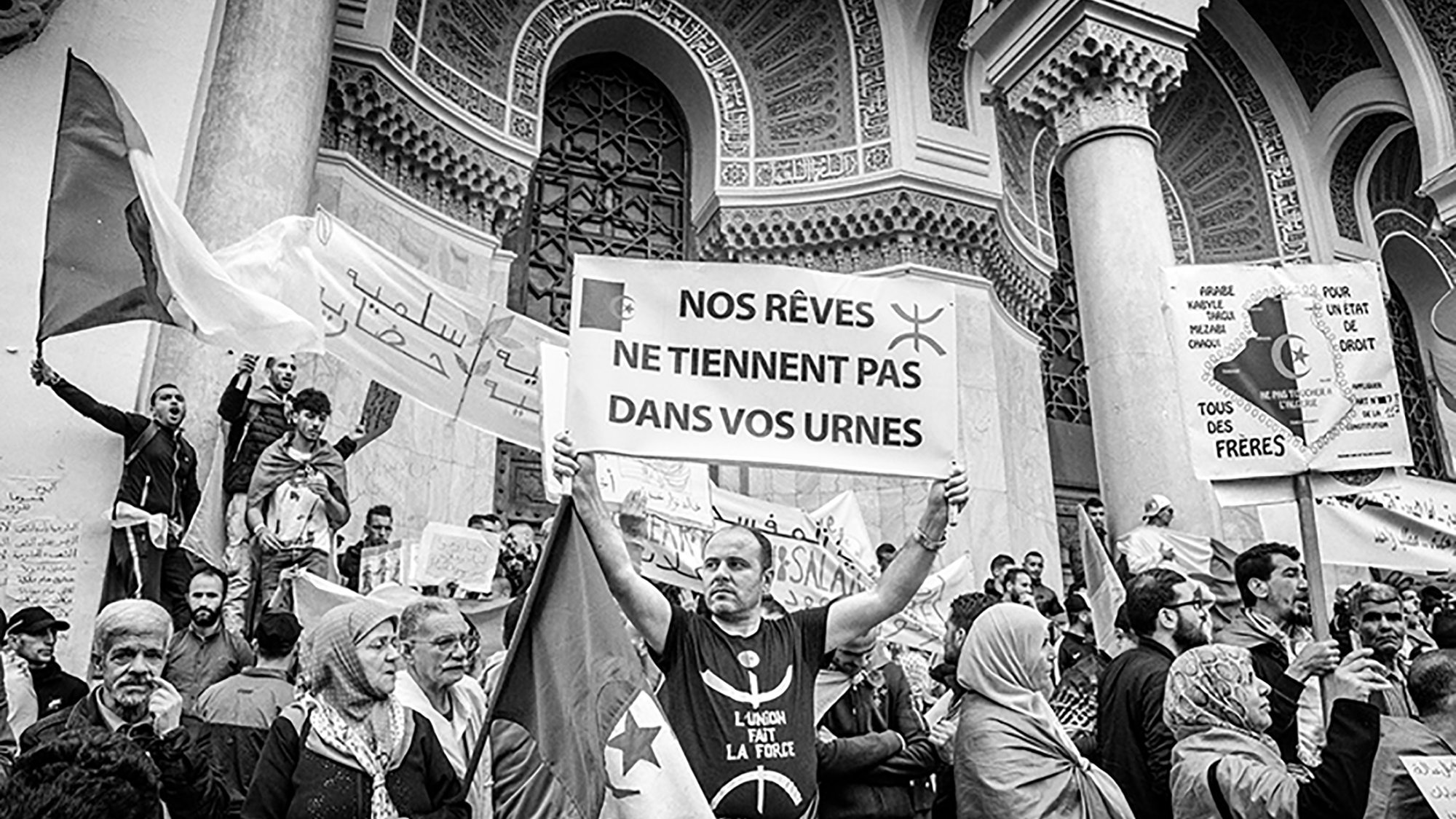 L'image montre une scène de manifestation où plusieurs personnes sont rassemblées. Au premier plan, un homme tient une grande pancarte qui affiche le message : "NOS RÊVES NE Tiennent PAS DANS VOS URNES". Les manifestants entourant cet homme expriment des sentiments de protestation, avec des drapeaux et des banderoles visibles. L'atmosphère semble chargée d'émotion et d'engagement politique, le tout dans un cadre architectural imposant. La photo est en noir et blanc, ce qui accentue l'intensité de la situation.
