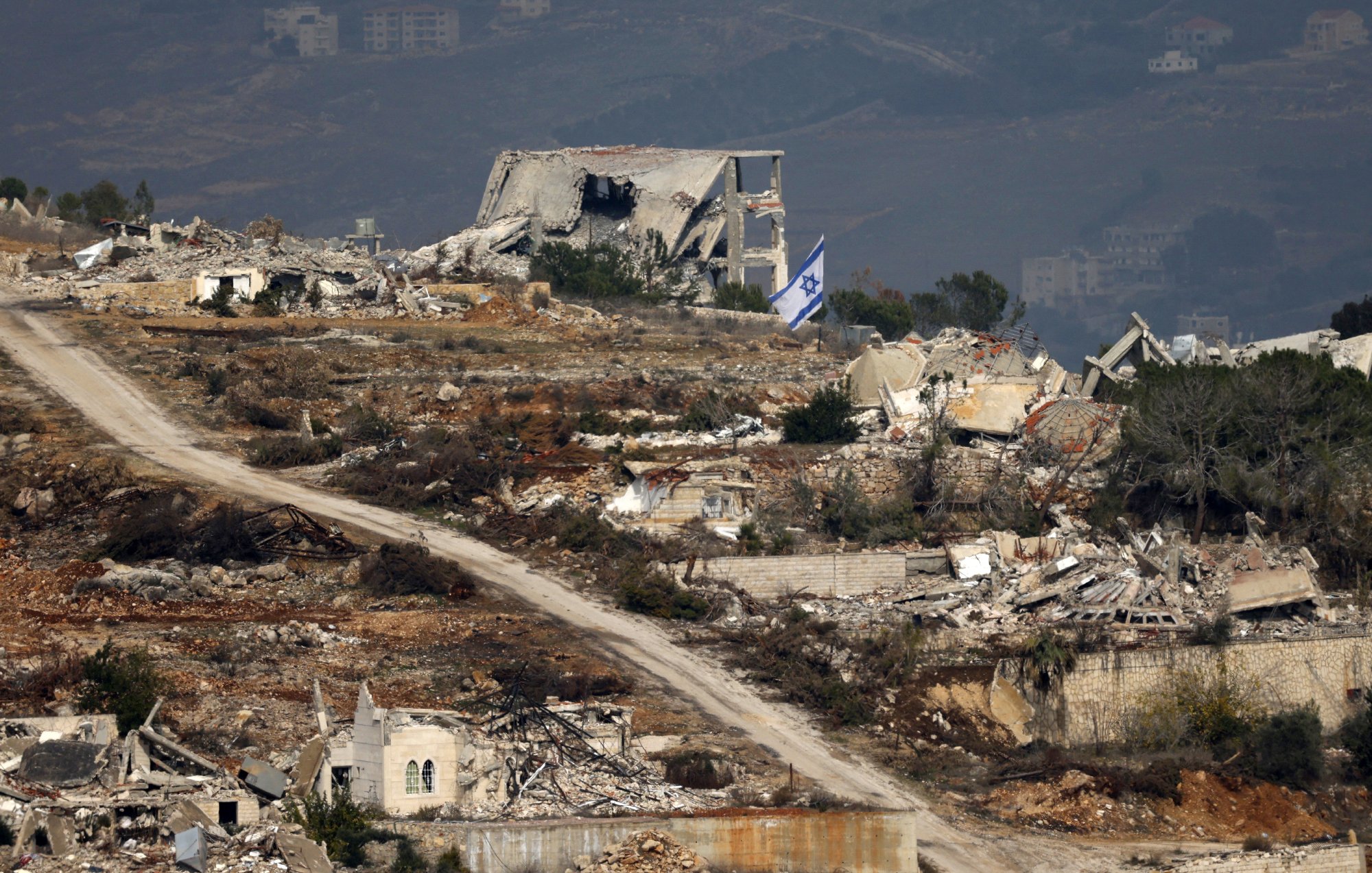 L'image montre un paysage de ruines, probablement dans une région touchée par un conflit. À l'arrière-plan, on peut voir des bâtiments détruits, avec des débris éparpillés. Une route de terre serpente à travers le paysage, et un drapeau israélien apparaît au premier plan, signalant la présence israélienne dans la zone. Le paysage est montagneux, avec des collines en arrière-plan, suggérant un environnement rural. L'image évoque une atmosphère de désolation et de conflit.