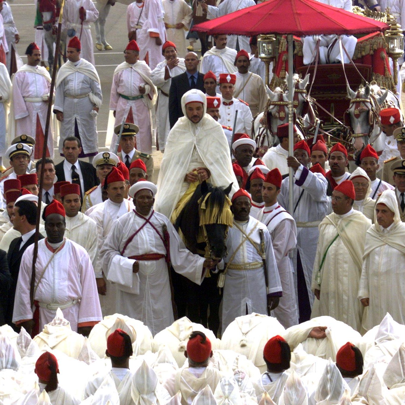 L'image montre un rassemblement cérémonial avec un grand nombre de personnes en tenue traditionnelle. Au centre, une figure importante est portée sur une plate-forme décorée, probablement un leader ou une personnalité éminente. Les participants portent des habits blancs et rouges, indiquant un événement culturel ou religieux. Des officiers en uniformes militaires se trouvent également parmi la foule, et certains photographient la scène. L'ambiance semble festive et respectueuse, typique d'une célébration ou d'une cérémonie officielle.