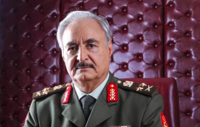 L'image montre un homme en tenue militaire, assis sur une chaise dans un cadre élégant. Il porte un uniforme avec des épaulettes ornées de médailles et d'insignes. L'arrière-plan est composé d'un panneau rembourré en cuir de couleur rouge, ce qui ajoute une touche de sophistication à l'ensemble. L'expression de l'homme est sérieuse et confiante.