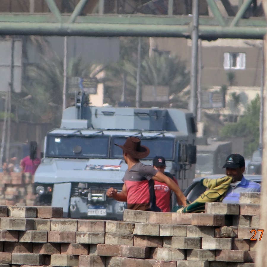 L'image montre une scène urbaine probablement marquée par des tensions. Au premier plan, des hommes sont vus près d'un empilement de briques, certains portant des vêtements de travail. En arrière-plan, un véhicule blindé est visible, probablement en patrouille ou en attente. L'environnement semble indiquer une zone de conflit ou de protestation, avec des bâtiments et des plantations à proximité. La date indiquée en bas de l'image (27 juillet 2013) peut faire allusion à un événement significatif dans le contexte de la photo.