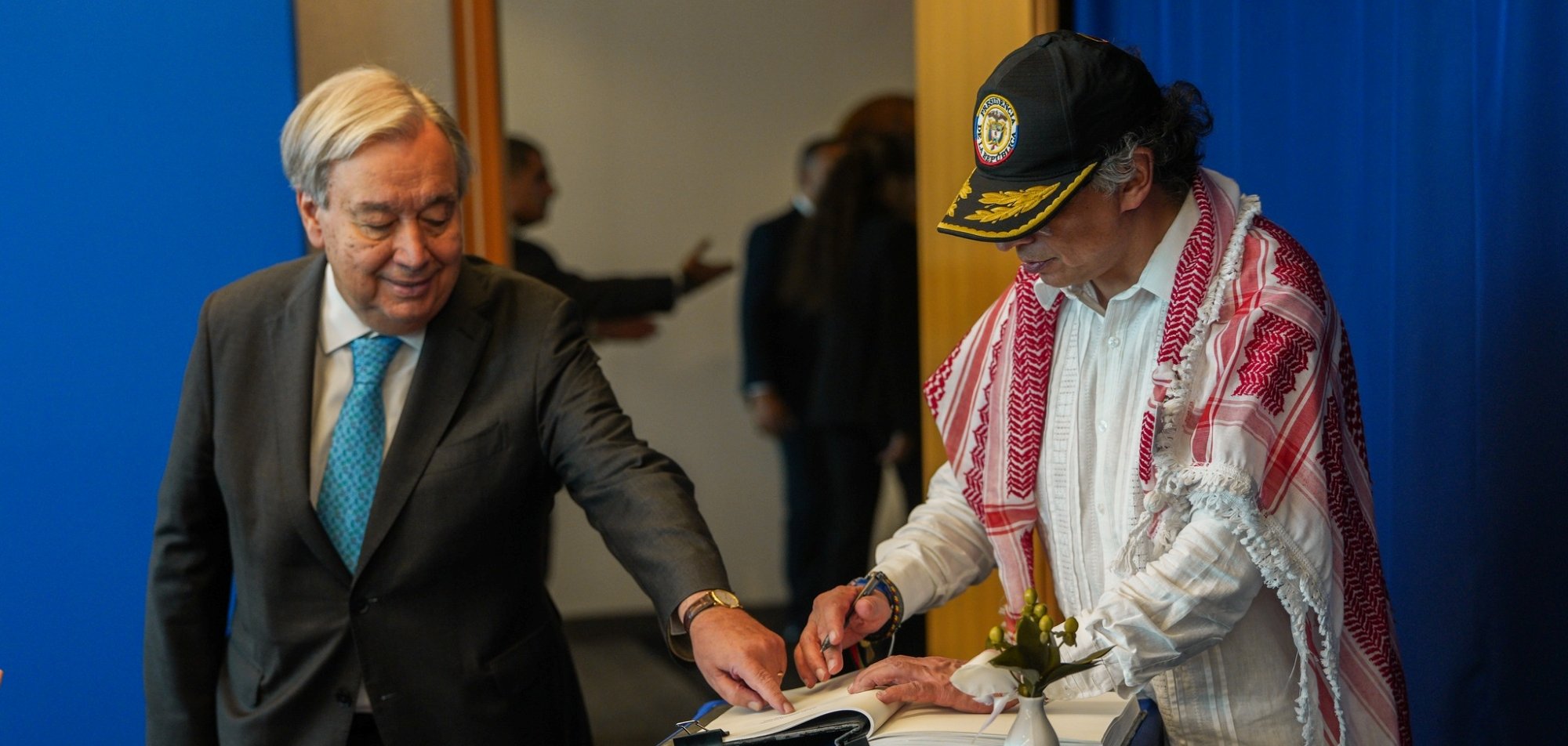 Two men are at a table; one is signing a book while the other observes. A blue backdrop is behind them.