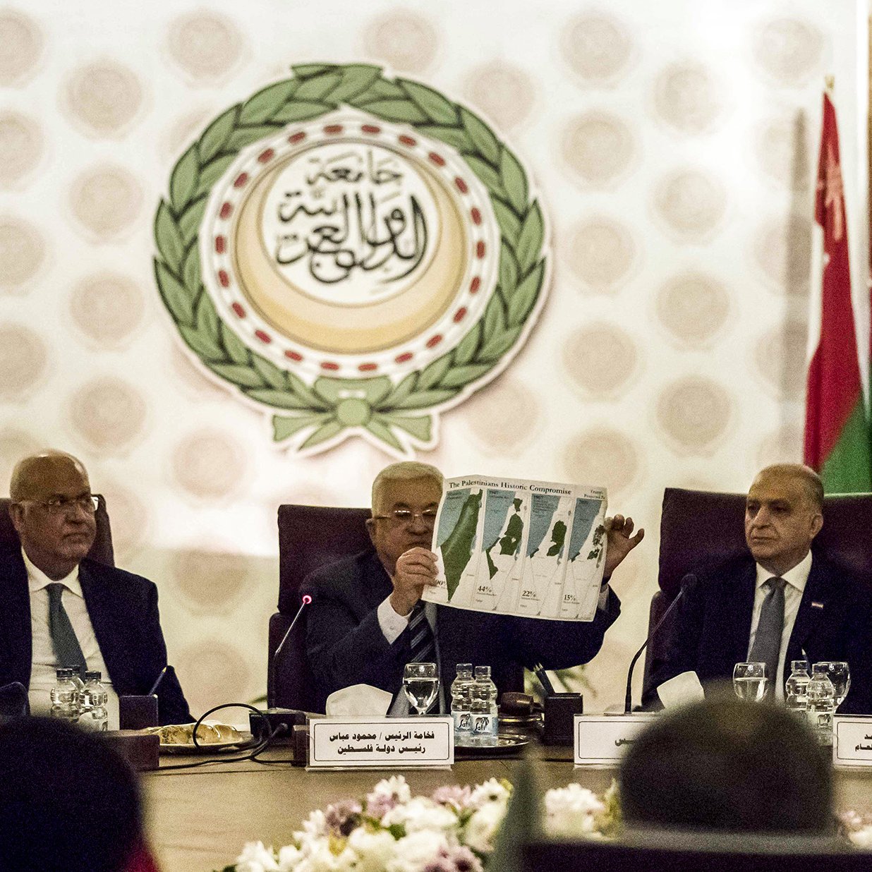 L'image montre une réunion officielle, probablement dans le cadre d'une organisation internationale. Plusieurs hommes sont assis autour d'une table dans un espace bien décoré, avec des drapeaux de différents pays en arrière-plan. L'un des hommes, au centre, tient un document ou un graphique qu'il montre aux autres participants. Les autres semblent attentifs et engagés dans la discussion. Les éléments visuels suggèrent une atmosphère sérieuse et formelle, en lien avec des discussions diplomatiques.