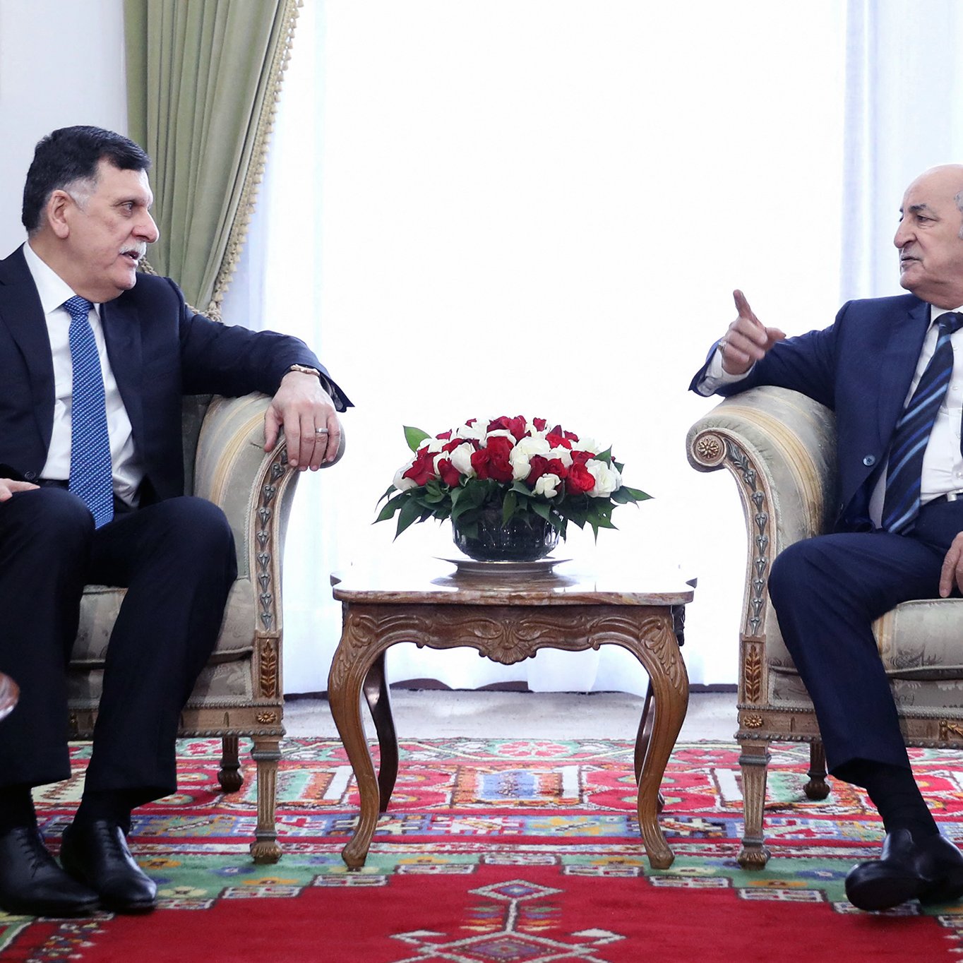 L'image montre deux hommes assis dans un environnement décoré de façon formelle. Ils sont en train de discuter, chacun semblant attentif à l'autre. Le premier homme, habillé en costume avec une cravate, se tourne légèrement vers le second. Ce dernier, également en costume, gesticule avec une main, ce qui pourrait indiquer qu'il est en train de faire un point ou de partager une idée. Entre eux, il y a une table basse ornée d'un arrangement floral. L'arrière-plan est lumineux et dégage une ambiance de rencontre officielle. Des éléments décoratifs, comme des rideaux et un tapis, ajoutent une touche élégante à la scène.