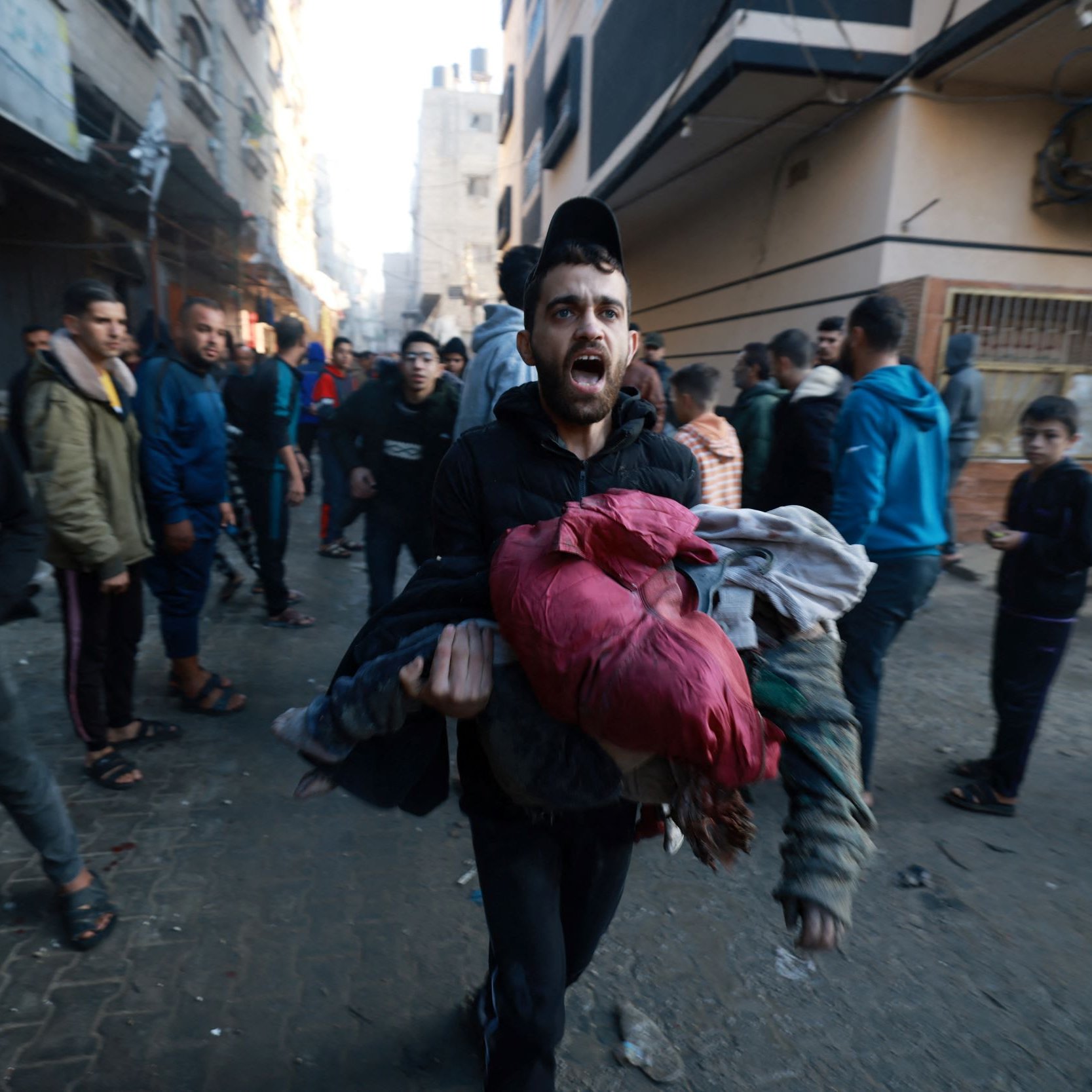 L'immagine mostra un gruppo di persone che si muovono in una strada affollata. Un uomo in primo piano, visibilmente agitato, sta correndo e tiene in braccio dei vestiti. Gli altri presenti nella scena sembrano essere coinvolti in una situazione di emergenza o caos, contribuendo a creare un'atmosfera di urgenza e tensione. Gli edifici circostanti appaiono deteriorati, suggerendo un contesto di conflitto o crisi.