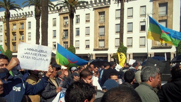 L'image montre une foule rassemblée lors d'une manifestation en plein air. On peut voir des gens tenant des pancartes et des drapeaux, dont certains affichent des couleurs vives, probablement liés à un mouvement culturel ou politique. Les bâtiments en arrière-plan semblent typiques d'un milieu urbain. L'ambiance semble être dynamique et engagée, reflétant les préoccupations des manifestants.