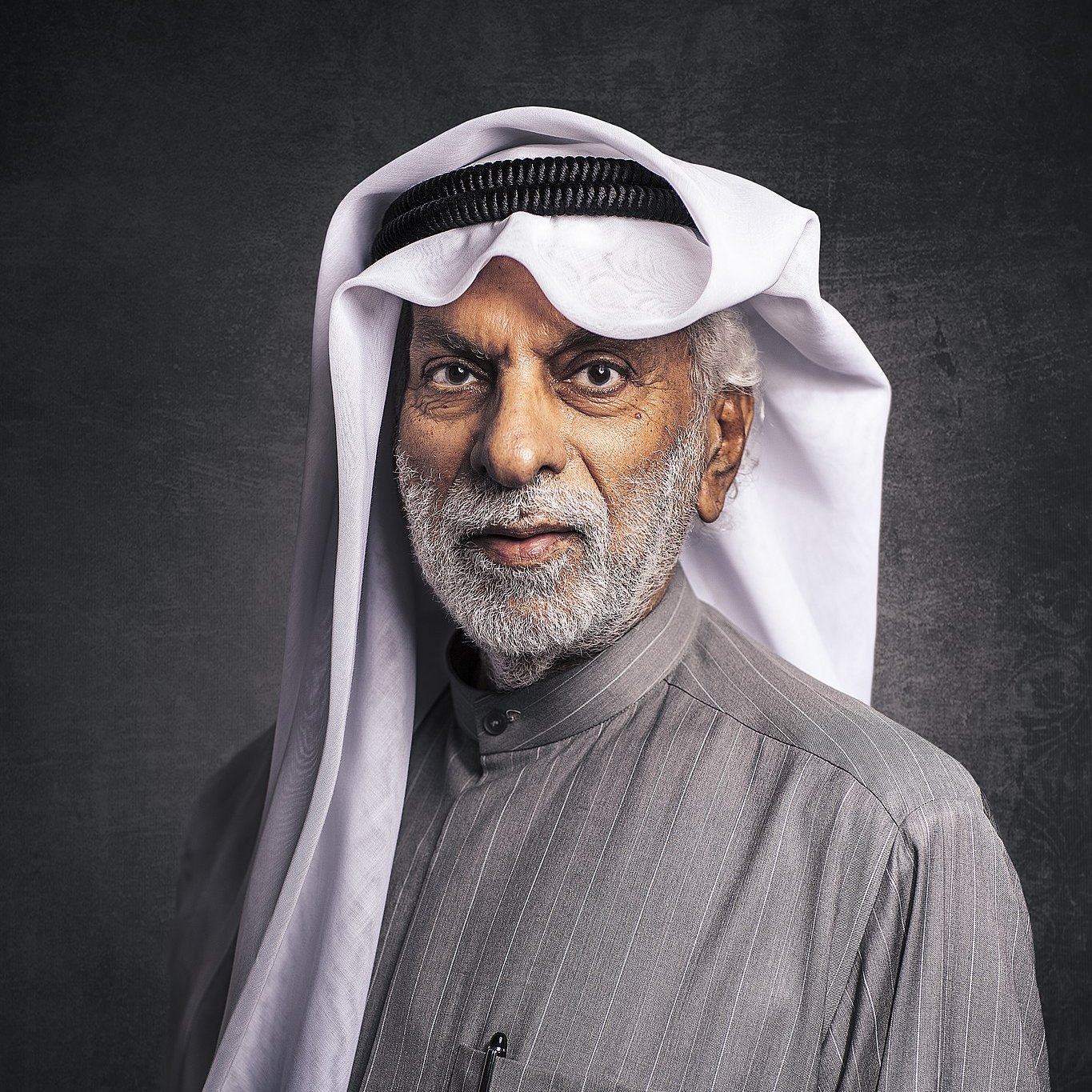 الصورة تظهر رجلاً مسنًا يرتدي الزي التقليدي العربي. لديه لحية رمادية وشعر أبيض، ويرتدي غترة بيضاء مع عقال أسود. يحمل ابتسامة لطيفة ويبدو أنه يتمتع بحضور مهيب. الخلفية مظلمة قليلاً مما يبرز تفاصيل وجهه وملابسه.