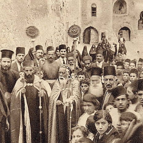 L'image montre un groupe de personnes rassemblées dans une ruelle étroite, probablement d'une ville historique. Au premier plan, on peut voir des hommes en habits traditionnels, certains portant des chapeaux typiques. Un homme central semble être un dignitaire religieux, accompagné d'une procession. En arrière-plan, des enfants et d'autres adultes regardent vers l'objectif, créant une atmosphère de communauté et de partage. Les bâtiments en pierre autour d'eux suggèrent un contexte ancien.