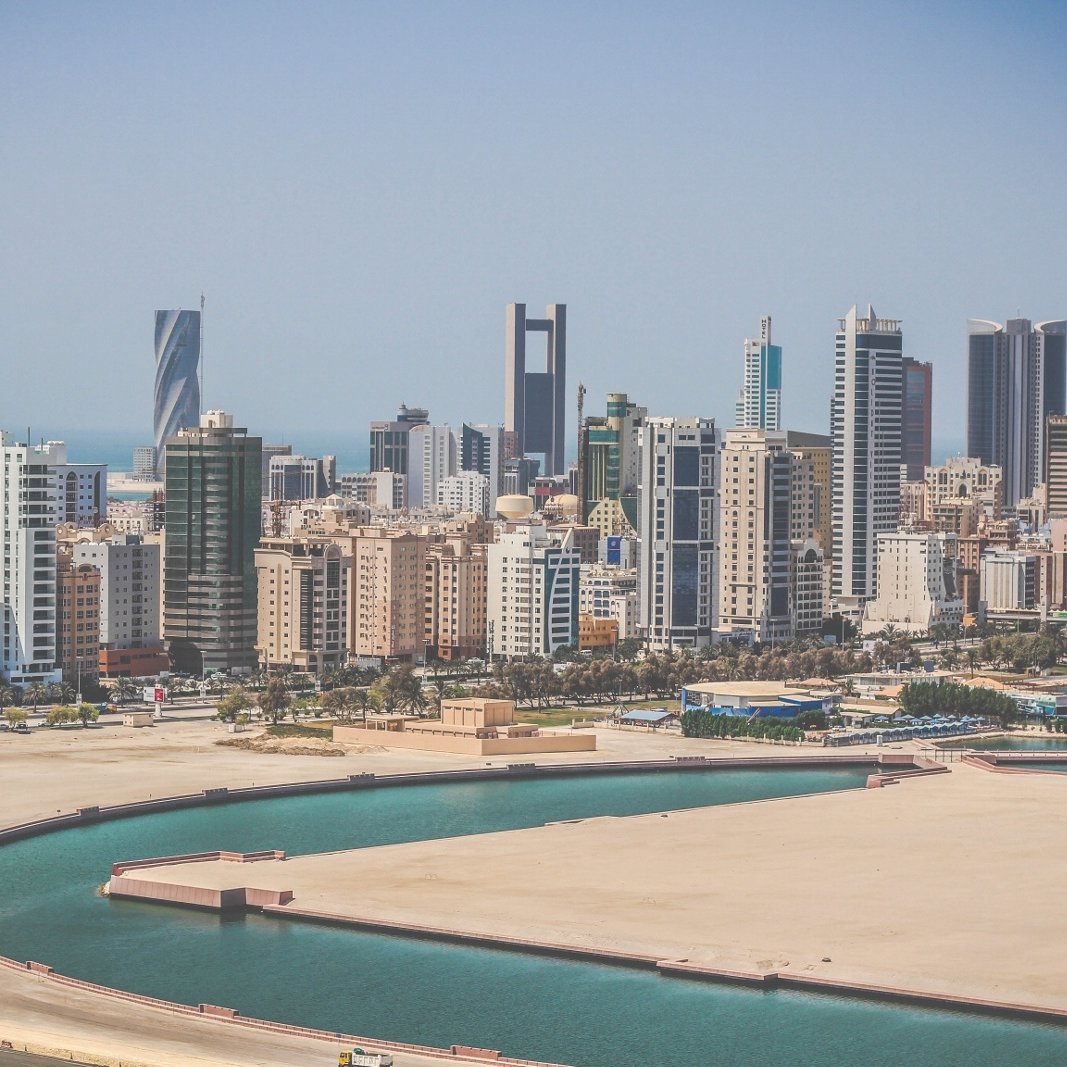 L'image montre un paysage urbain moderne avec une vue sur une ville côtière. On peut voir de nombreux immeubles de grande hauteur, représentant une architecture contemporaine, avec des designs variés allant des structures angulaires aux façades en verre. À l'arrière-plan, la mer est visible, ainsi qu'une baie ou un port. Le ciel est clair et ensoleillé, ce qui donne une impression de chaleur et de vivacité à la scène. La composition évoque une ambiance urbaine dynamique, typique de villes en développement.