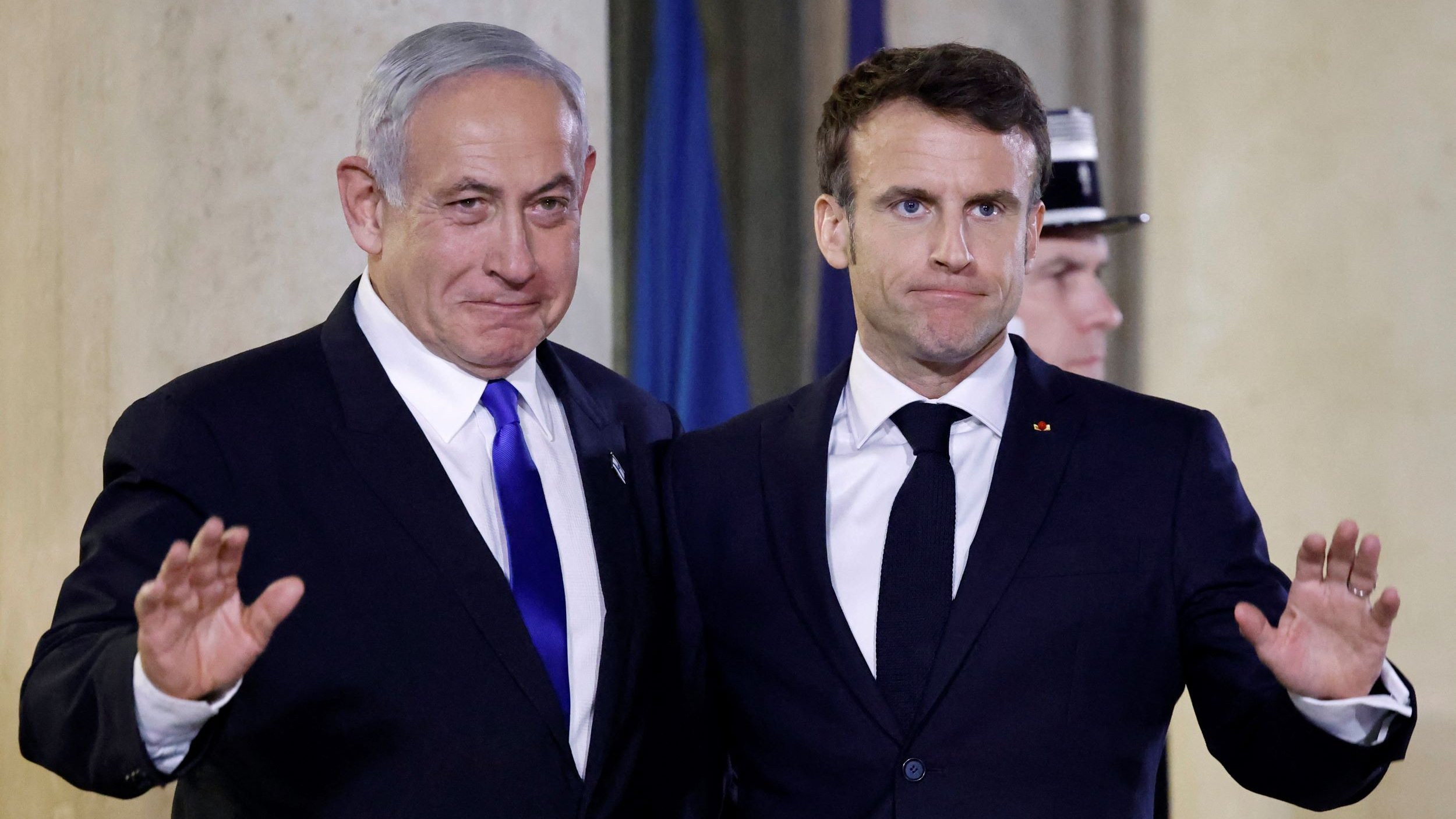 الصورة تظهر شخصين يقفان معًا أمام خلفية مميزة. يبدو أن أحدهما يرتدي بدلة زرقاء وقميص أبيض مع ربطة عنق زرقاء، بينما الآخر يرتدي بدلة سوداء وقميص أبيض مع ربطة عنق داكنة. كلاهما يبتسم قليلاً ويرفعان أيديهما في إشارة ودية. الجو العام في الصورة يبدو رسميًا، مما يشير إلى أن اللقاء قد يكون في مناسبة دبلوماسية أو رسمية.