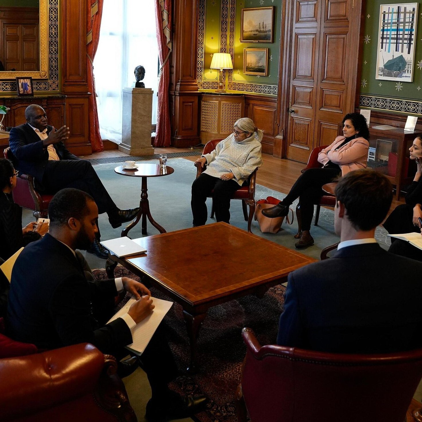 Nell'immagine si vede un incontro formale all'interno di una stanza ben arredata, con pareti in legno e decorazioni artistiche. Un gruppo di persone è riunito attorno a un tavolino centrale. Alcuni partecipanti sono seduti su poltrone, mentre altri sono seduti su uno sgabello o in posizioni più informali. Alcuni hanno in mano blocchi note o penne, suggerendo che stanno prendendo appunti. La luce naturale entra attraverso le finestre, creando un'atmosfera accogliente. Sullo sfondo, si intravedono alcune opere d'arte e un camino, che aggiungono un tocco elegante all'ambiente.