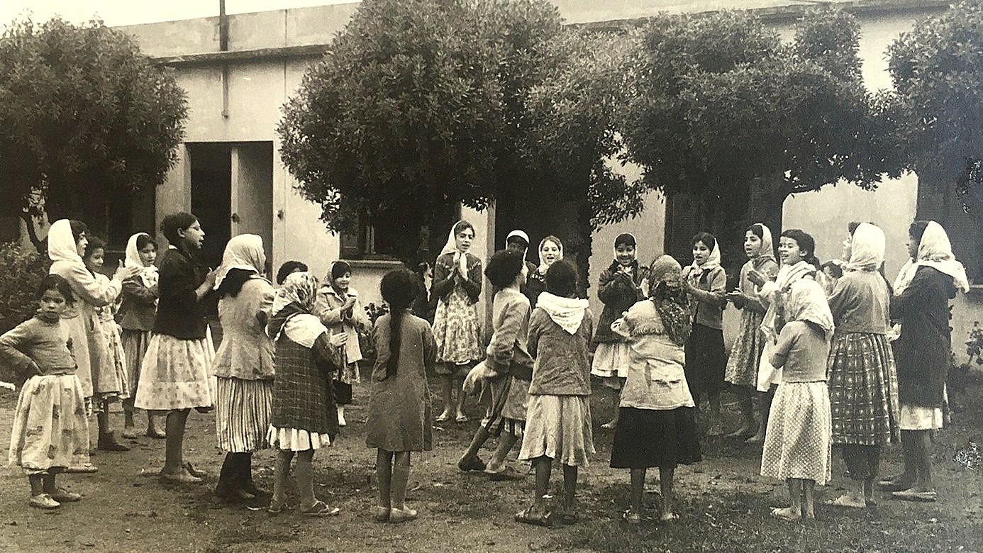 تظهر في الصورة مجموعة من الفتيات والأطفال يلعبون في ساحة مفتوحة. يبدو أنهن يرتدين ملابس تقليدية، وبعضهن يحمل غطاء رأس. الفتيات محاطة بأشجار، مما يضفي جوًا طبيعيًا على المكان. يبدون مشغولات باللعب والضحك، في مشهد يعكس روح الطفولة والنشاط. الخلفية بسيطة، مع وجود مبنى مكون من جدران بسيطة.