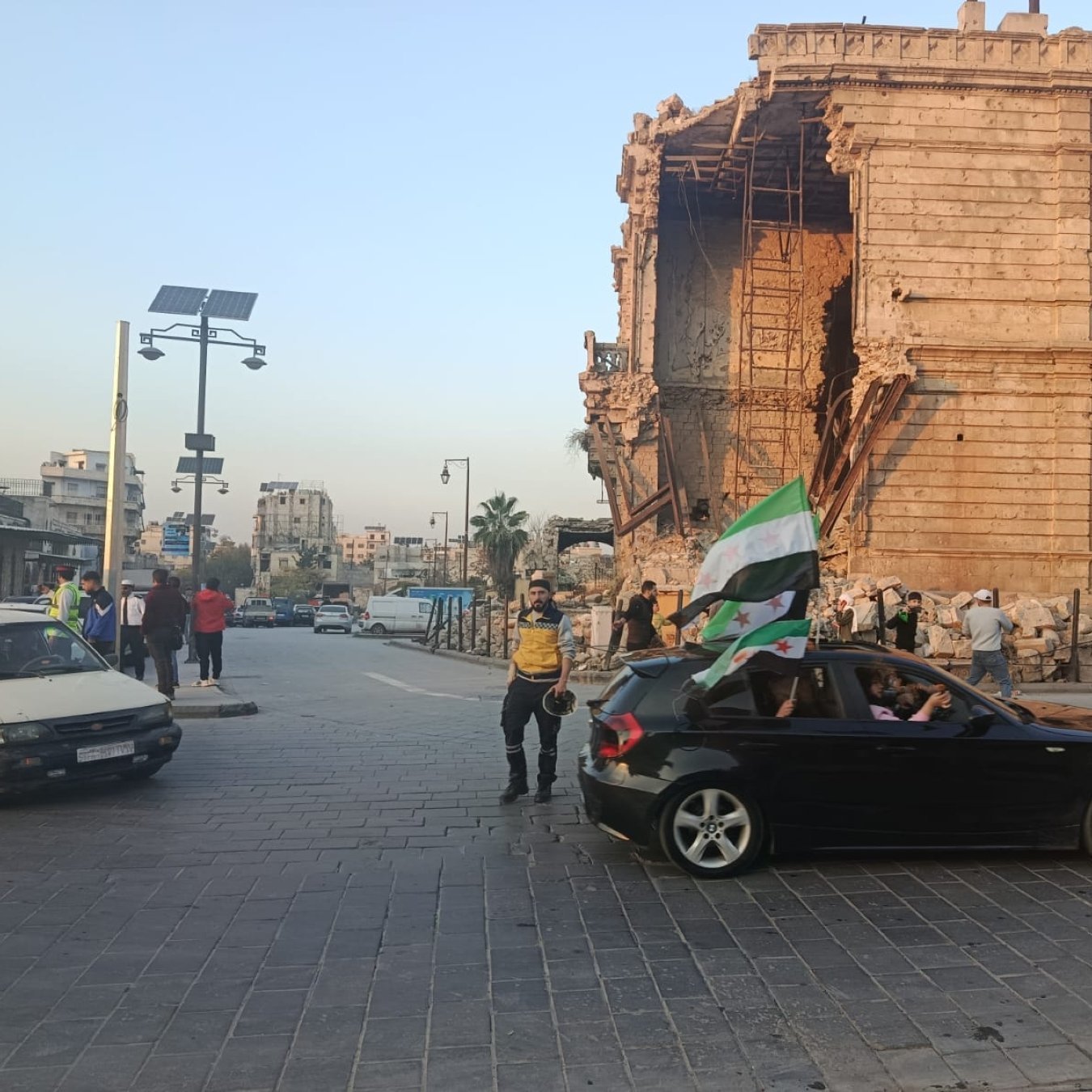 Una strada con una costruzione danneggiata, persone e un'auto con una bandiera.