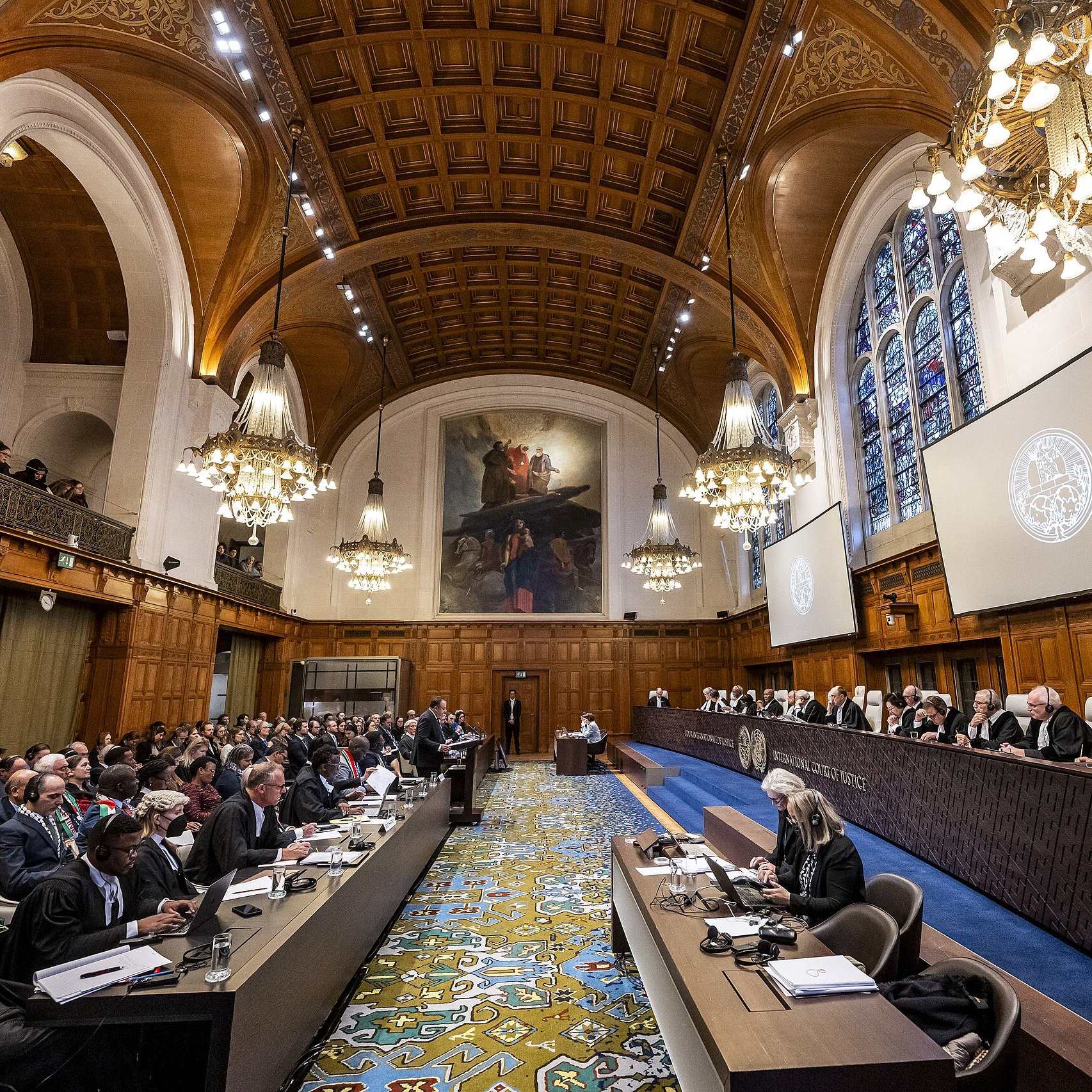 The image depicts an interior of a grand courtroom, likely in an international court such as the International Court of Justice. The room features high wooden ceilings with intricate designs and ornate chandeliers providing warm lighting. Large windows with stained glass allow natural light to filter in. In the foreground, there are rows of judges seated on a raised platform, alongside legal representatives and observers. The layout includes a long table with officials on one side and a broad audience on the other. The design of the room is both formal and historic, emphasizing the importance of the proceedings taking place.
