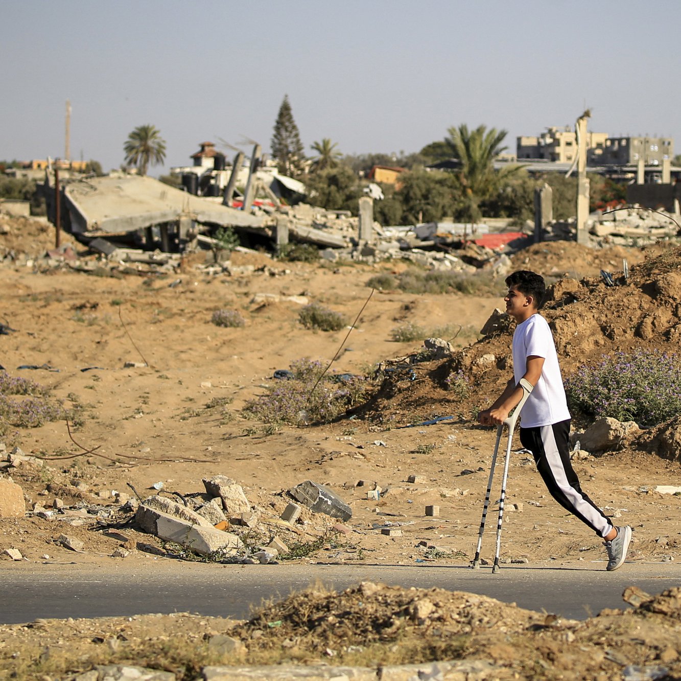 L'immagine mostra un giovane che cammina con le stampelle in un'area distrutta, circondata da macerie e detriti. Sullo sfondo si possono vedere edifici in rovina e un paesaggio desolato, con sabbia e vegetazione sparsa. La scena trasmette un senso di difficoltà e resilienza in un contesto di devastazione.