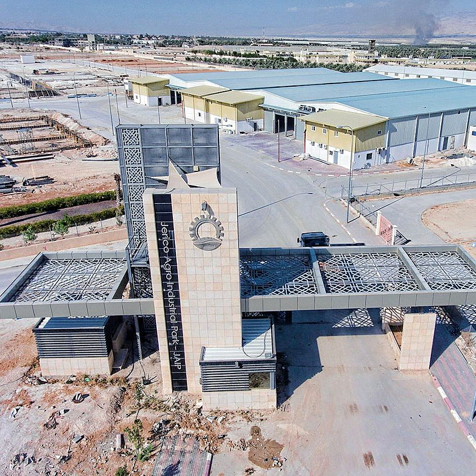 L'image montre un complexe industriel ou commercial avec une entrée bien marquée. On peut voir un portail moderne, probablement en métal et en pierre, qui semble être la pièce maîtresse de l'entrée. À l'arrière plan, il y a plusieurs bâtiments de différentes tailles, dont un plus grand qui pourrait servir de site de production ou d'entrepôt. Le terrain environnant est désertique,avec quelques infrastructures comme des panneaux solaires. En arrière-plan, on distingue un nuage de fumée, suggérant une activité ou un incendie à proximité.