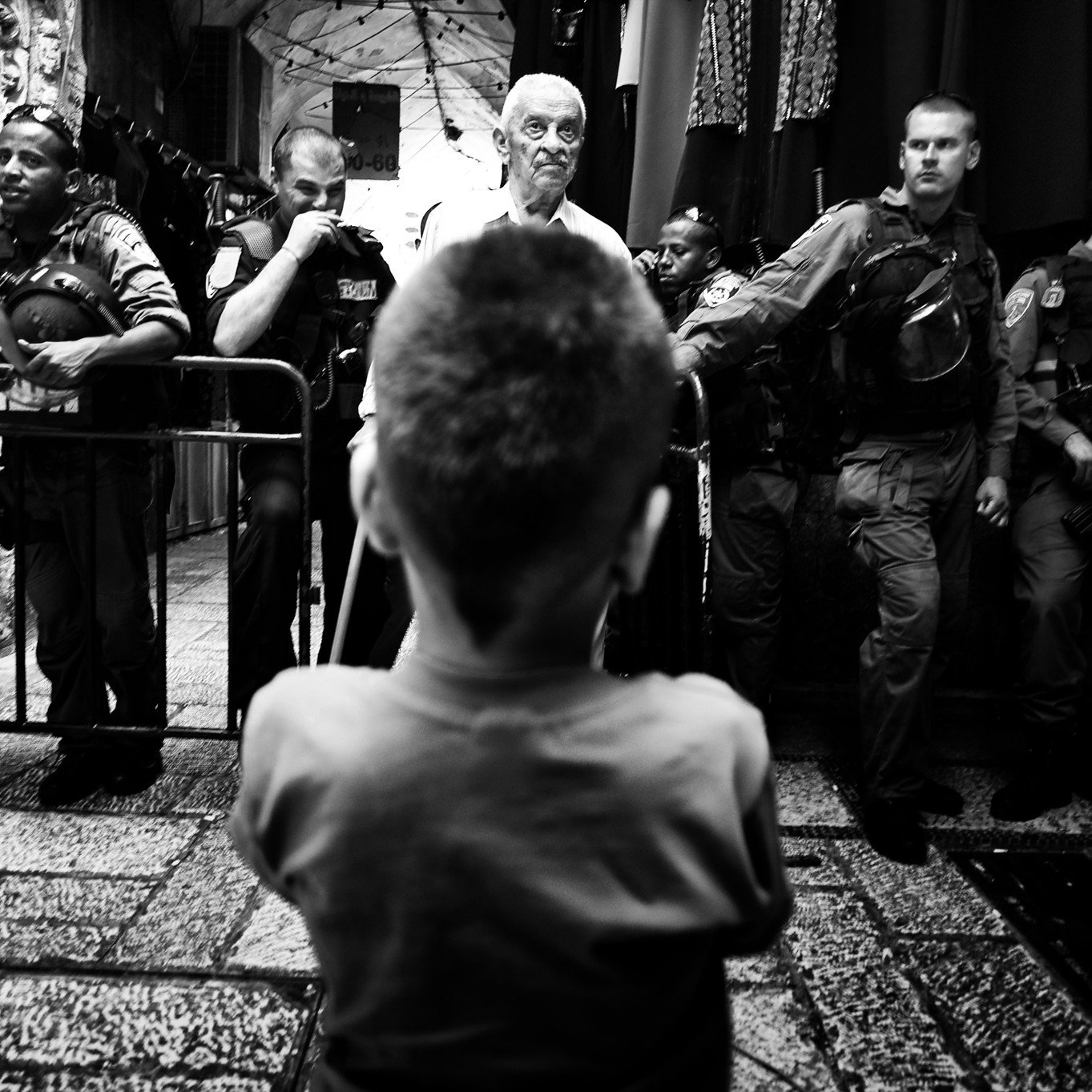 L'image montre un enfant vu de l'arrière, regardant vers un groupe de personnes, dont certaines semblent être des policiers ou des soldats. L'atmosphère est tendue, avec des barrières de sécurité en place et une composition en noir et blanc qui accentue le contraste entre les figures. L'enfant, central dans la scène, semble intrigué ou préoccupé par la situation qui se déroule devant lui. Les autres personnes sont alignées, probablement en position d'intervention.