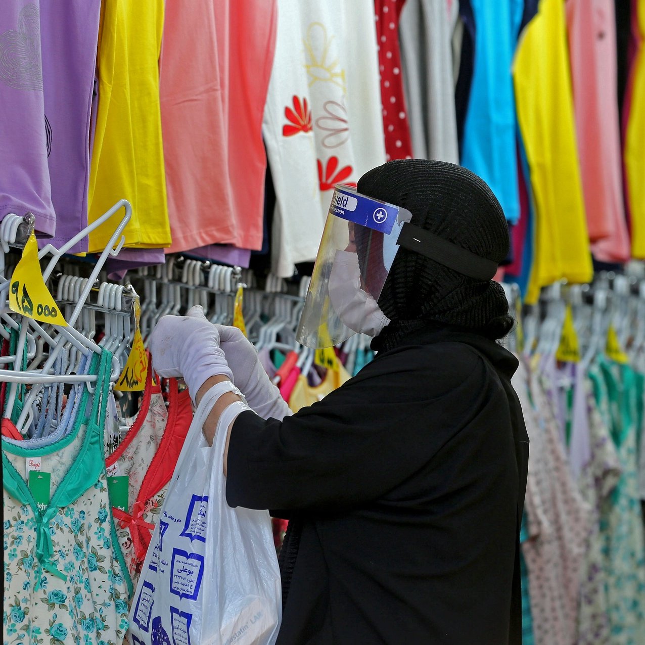 L'image montre une personne dans un magasin de vêtements, portant un hijab et un écran facial. Elle porte des gants et examine des vêtements accrochés sur des cintres. Les étagères à proximité sont remplies de vêtements colorés, principalement des t-shirts et des leggings, dans des teintes vives comme le violet, le jaune et le vert. L'ambiance semble être celle d'un environnement de shopping où l'on cherche des articles vestimentaires.