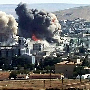 L'image montre un paysage urbain après une explosion, avec d'importants nuages de fumée et des décombres. Des bâtiments sont partiellement détruits, et l'atmosphère est sombre en raison de la fumée épaisse. Le sol semble être rural, avec des champs et quelques structures en arrière-plan. Cette scène évoque des conséquences dramatiques d'un conflit ou d'une attaque.