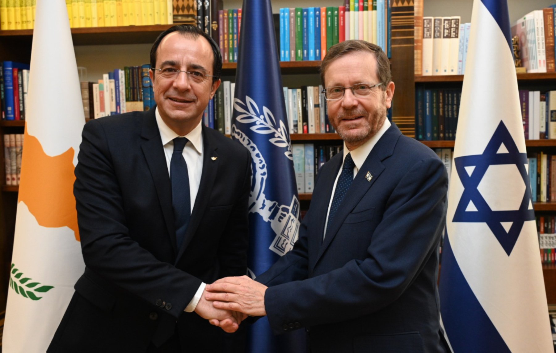 Deux hommes en costume se serrent la main devant des drapeaux et des étagères de livres.