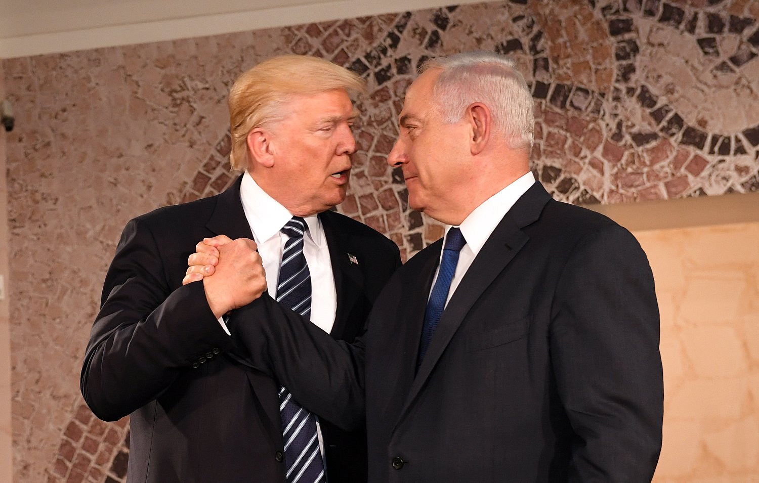 L'image montre deux hommes en train de se serrer la main avec des expressions sérieuses. Ils sont habillés en costume, l'un portant une cravate rayée et l'autre une cravate unie. L'arrière-plan possède des motifs décoratifs. Cette scène semble se dérouler lors d'un événement officiel ou d'une rencontre diplomatique.