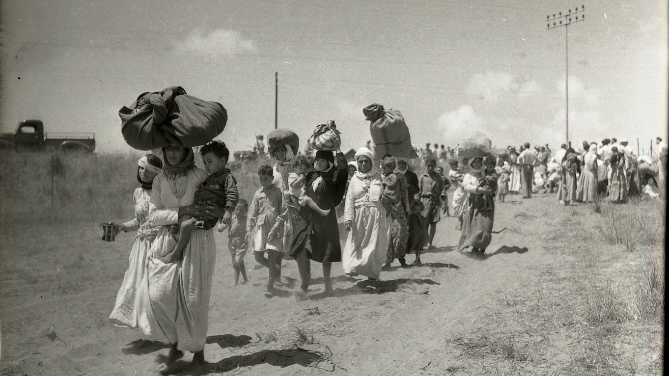 L'image montre un groupe de personnes marchant le long d'un chemin poussiéreux. Certaines femmes portent de lourdes charges sur la tête, tandis que d'autres tiennent des enfants par la main. La scène semble illustrer un moment de déplacement ou de migration, avec des paysages arides en arrière-plan. Des nuages blancs ponctuent le ciel, et on peut voir des lignes électriques au loin. L'atmosphère de la photo évoque la lutte et la détermination des personnes en marche.