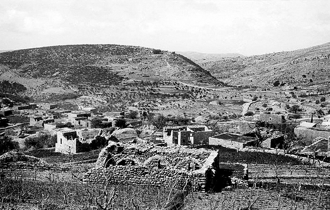 L'image montre un paysage rural en noir et blanc, où l'on peut apercevoir des ruines de structures en pierre, témoignant d'une ancienne habitation. Le terrain est vallonné, avec des collines en arrière-plan et une végétation clairsemée. Certaines zones semblent cultivées ou utilisées pour l'élevage, tandis que d'autres présentent des traces de dégradation ou d'abandon. L'atmosphère générale est paisible, mais elle évoque également une histoire de déclin ou de dévastation.