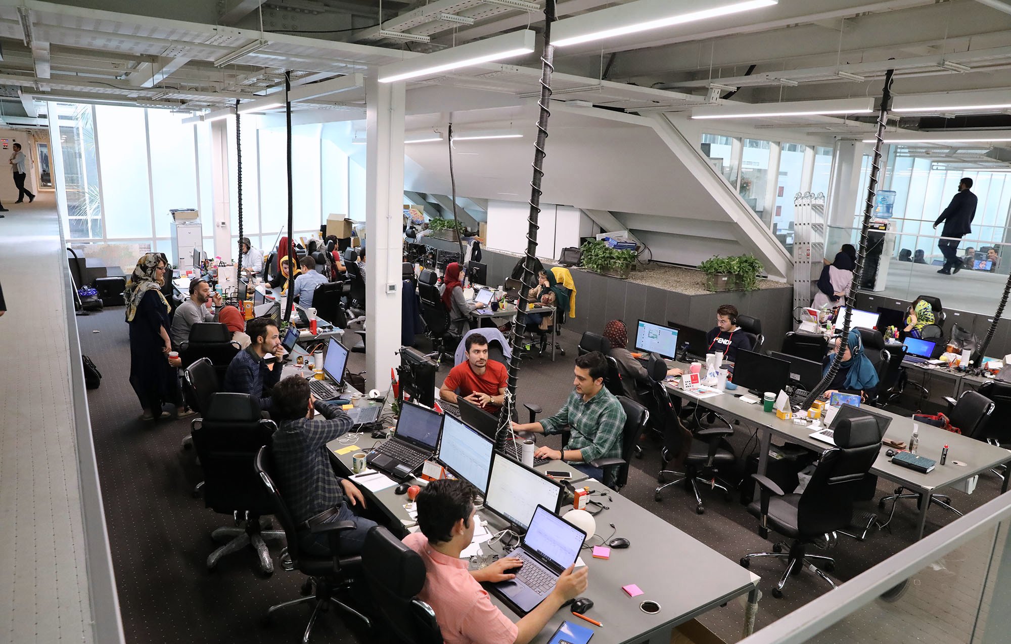 L'image montre un espace de bureau moderne et dynamique. Il y a plusieurs personnes qui travaillent à des bureaux équipés d'ordinateurs. L'environnement semble collaboratif, avec des employés assis autour de tables, certains en train de discuter, tandis que d'autres se concentrent sur leur travail. Les bureaux sont bien éclairés et décorés avec des plantes. On peut voir des objets personnels et des collations éparpillés sur les tables, ce qui donne une atmosphère informelle et conviviale.