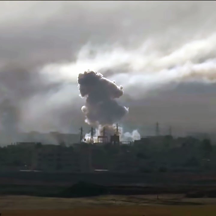 L'image montre une scène dramatique avec des nuages de fumée épais s'élevant dans le ciel, probablement à la suite d'une explosion ou d'un bombardement. On peut voir des silhouettes d'infrastructures industrielles ou urbaines partiellement obscurcies par la fumée. L'atmosphère est sombre et chargée, suggérant des conditions de conflit ou de destruction. La lumière semble faible, ajoutant à l'impression de chaos et de détresse.